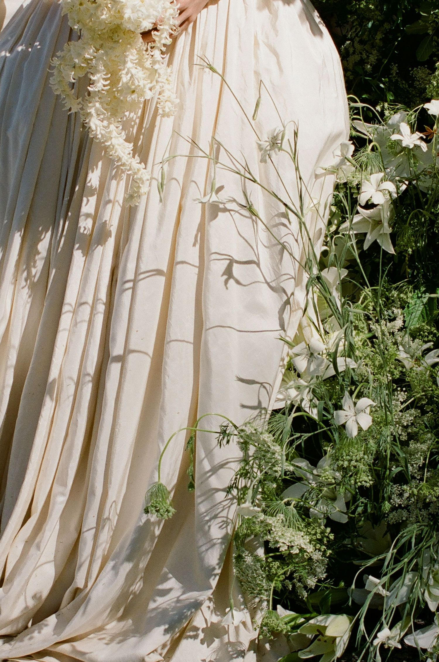 Bride in flowing ivory gown holding white floral bouquet beside lush green garden flowers in soft natural light.