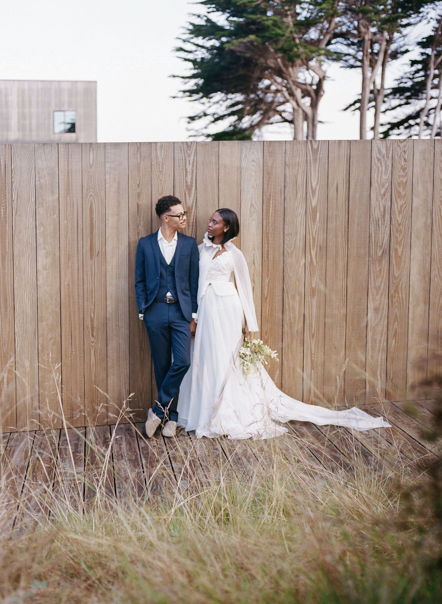 Couple in wedding attire standing against wooden fence, bride in white gown with bouquet, groom in navy suit.