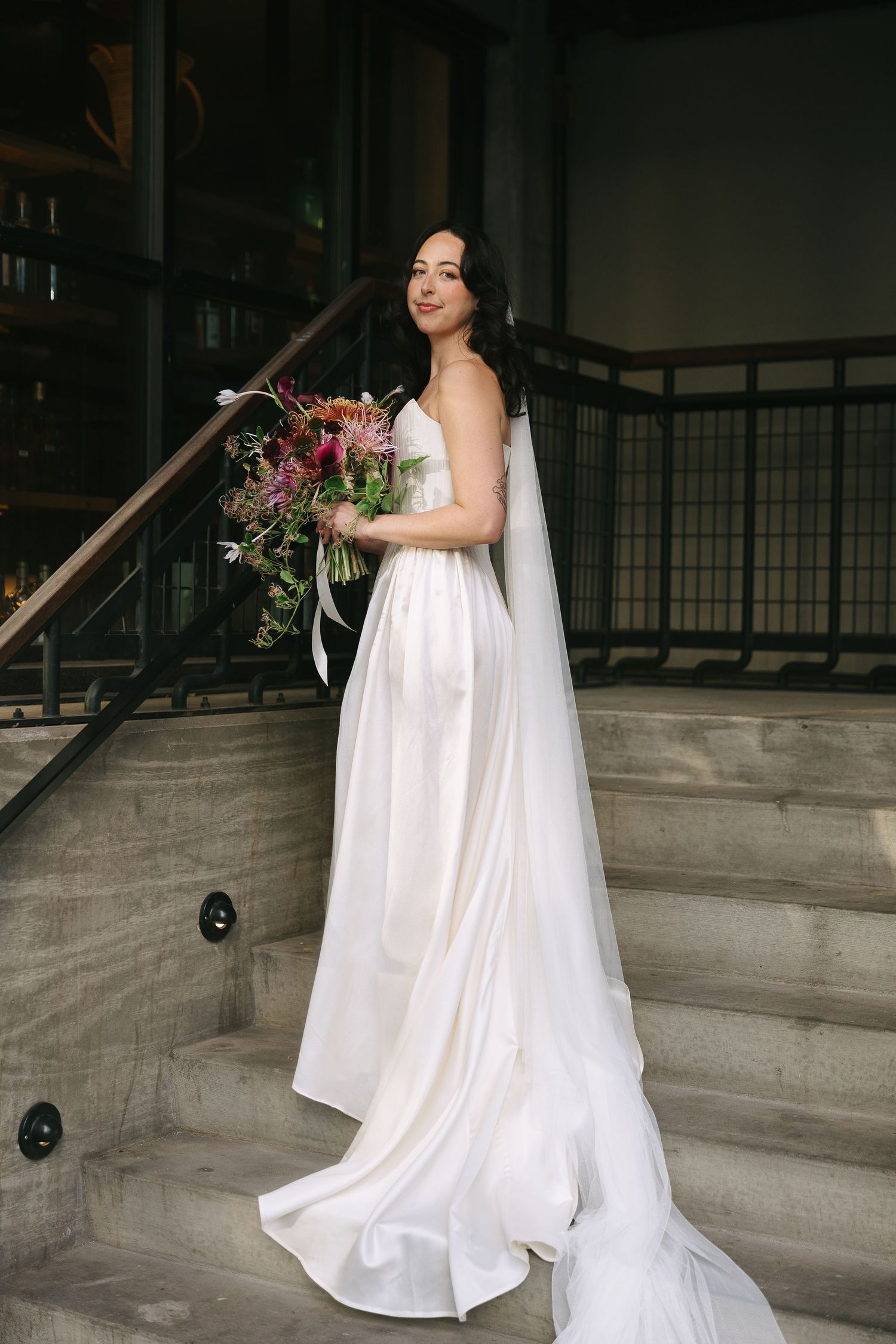 A bride in a flowing white gown stands on concrete stairs holding a dramatic fall-colored bouquet.