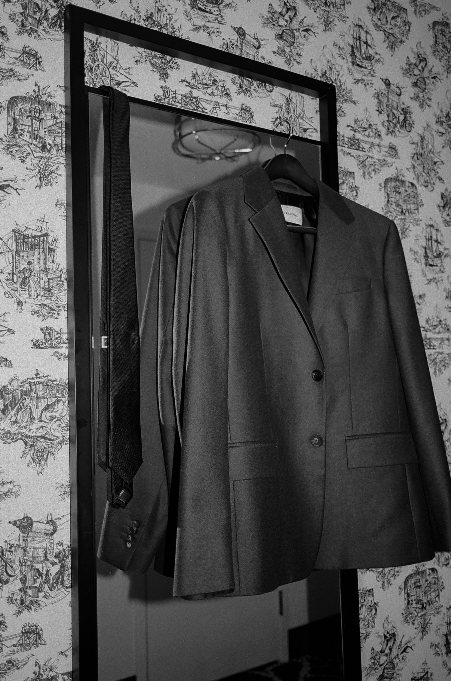 A black and white photograph of a formal suit jacket hanging on a mirror against floral wallpaper.