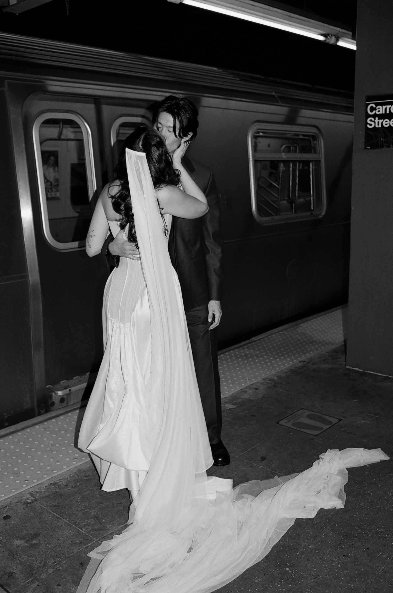A bride in a flowing white gown walks along a subway platform with her train trailing behind her in black and white.