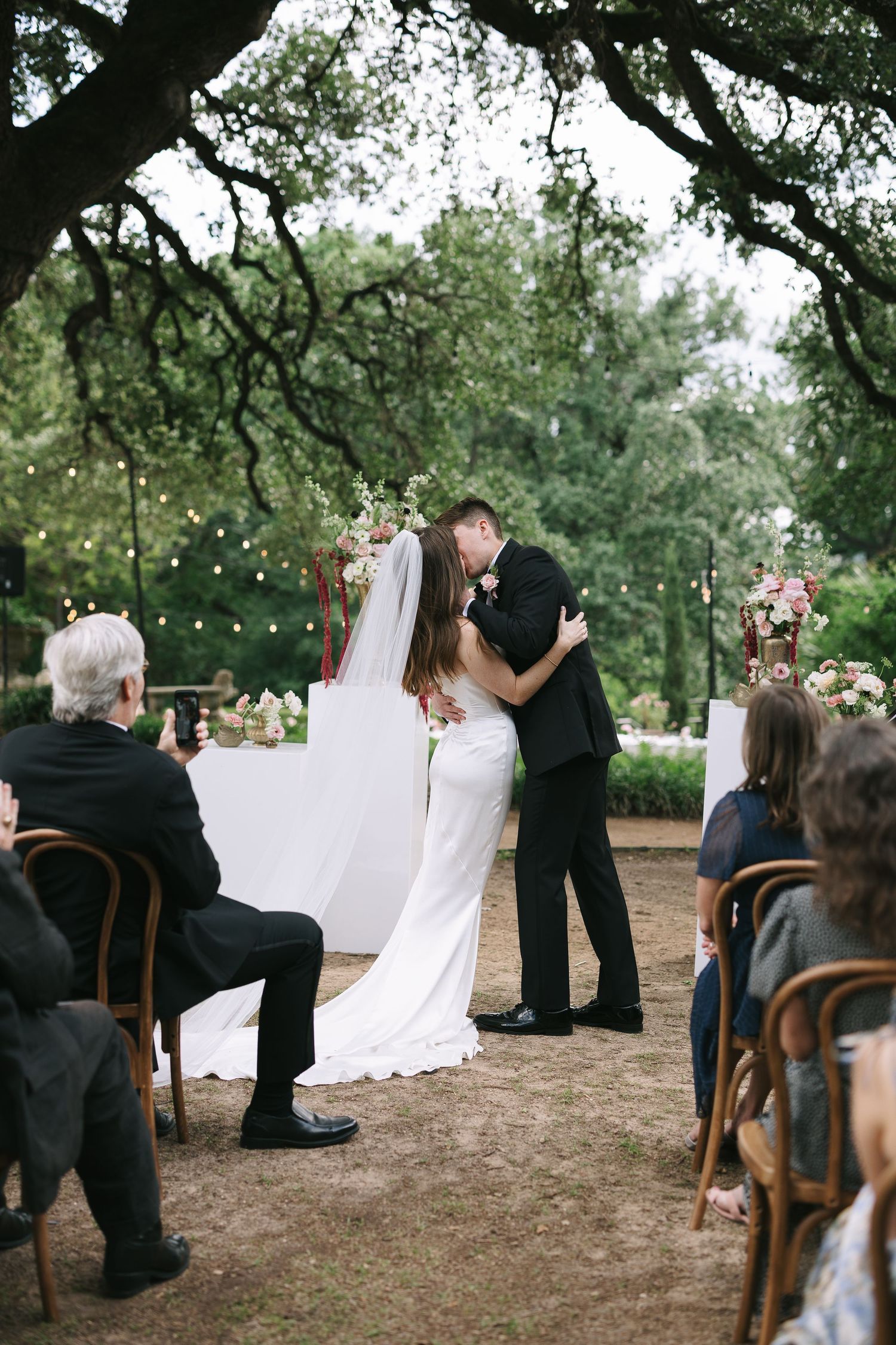 Intimate outdoor wedding ceremony with white backdrop and string lights under oak tree canopy.