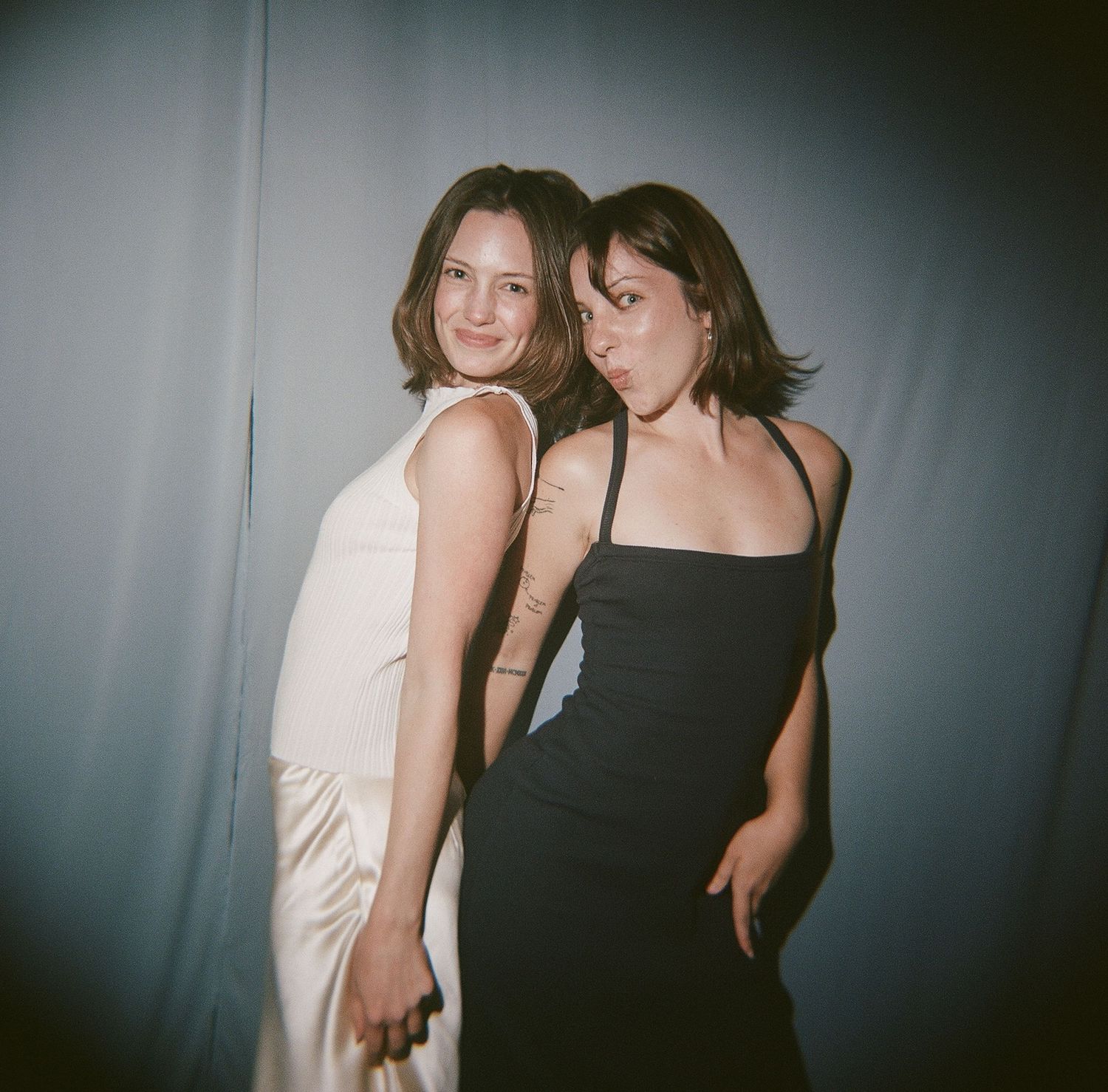 Two young women posing together, one in a white dress and one in a black dress, against a gray backdrop.
