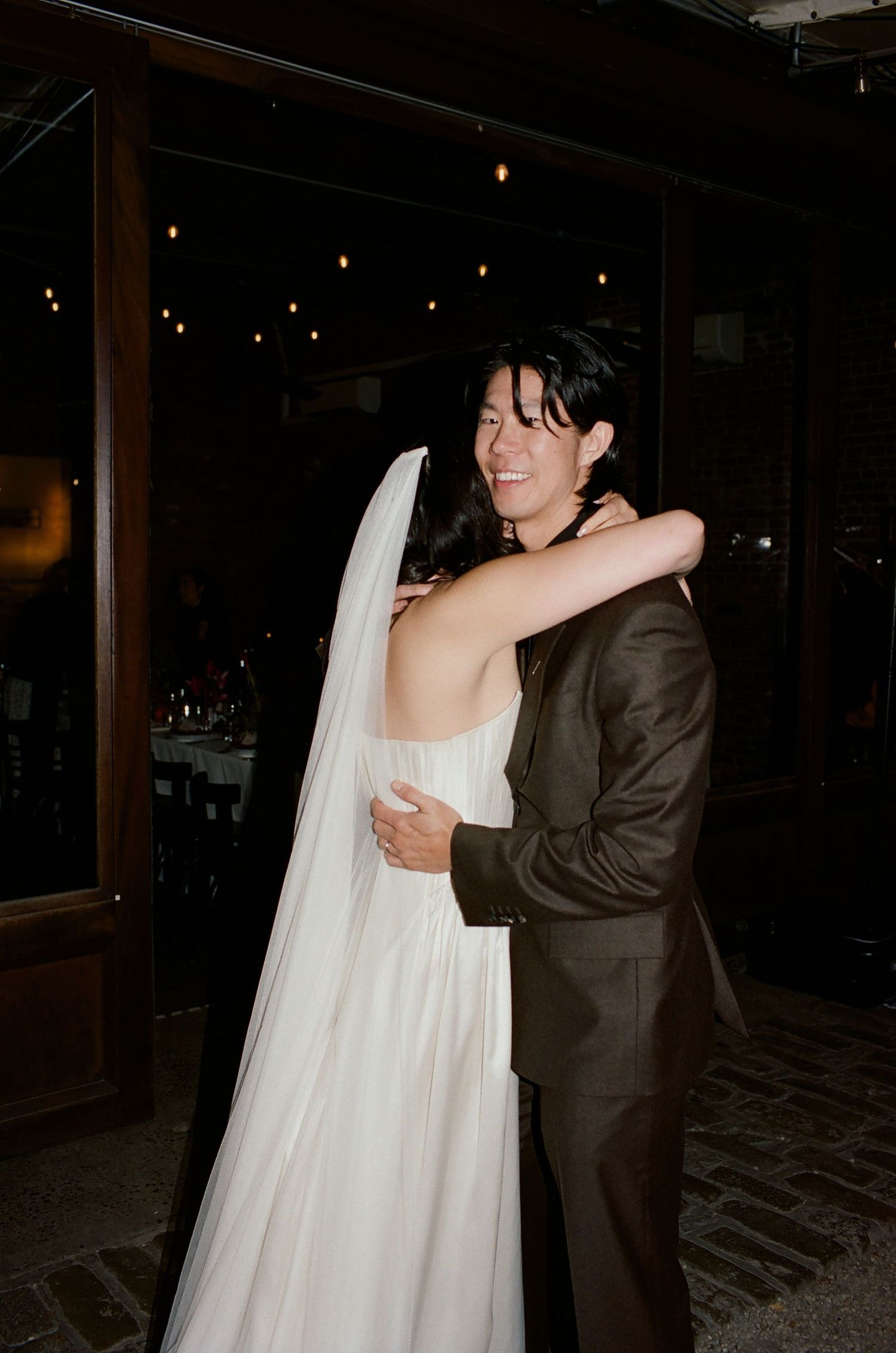 A couple embraces during their wedding celebration at night with string lights glowing in the background.