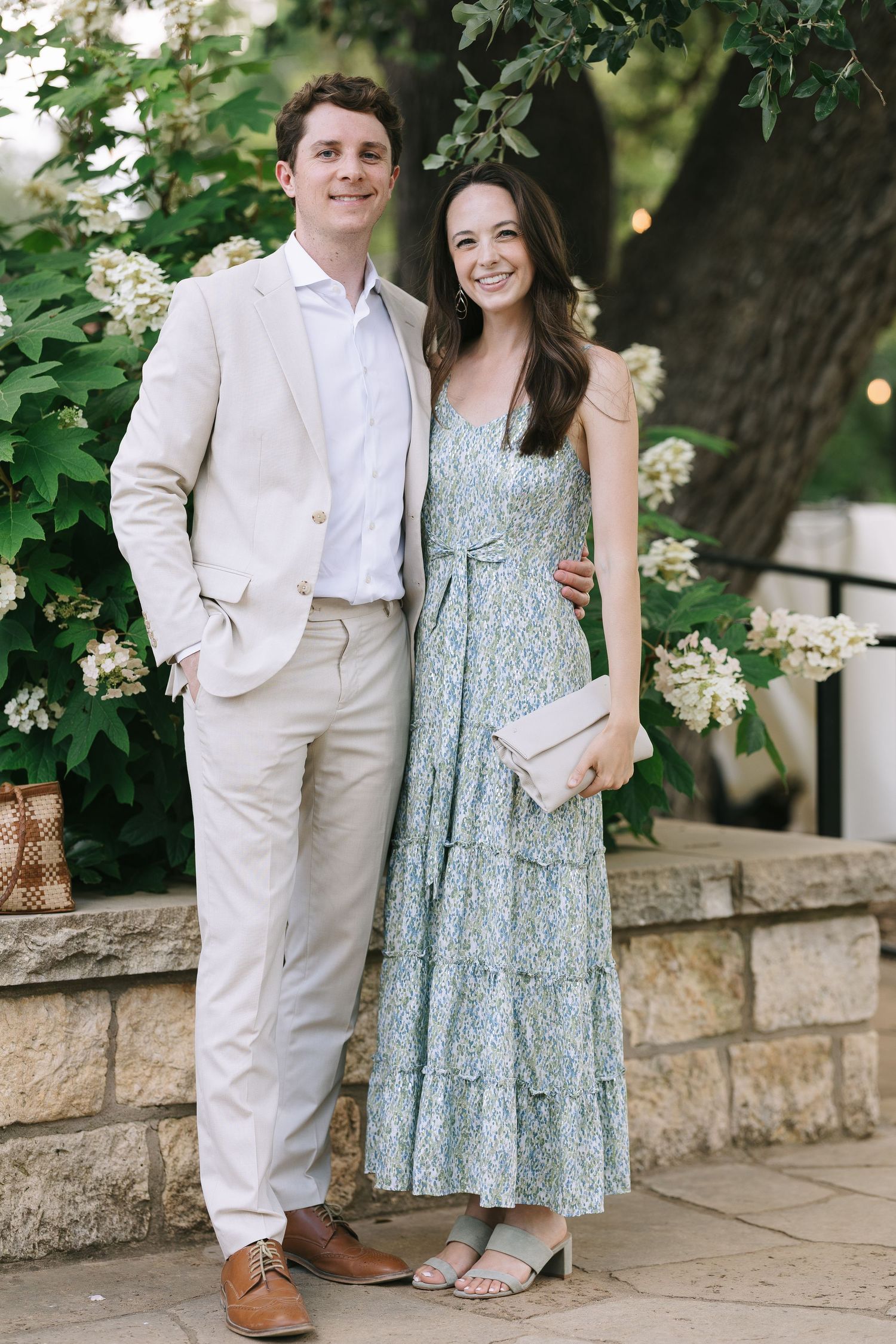 A couple poses together outdoors against a stone wall with greenery, wearing summer attire.
