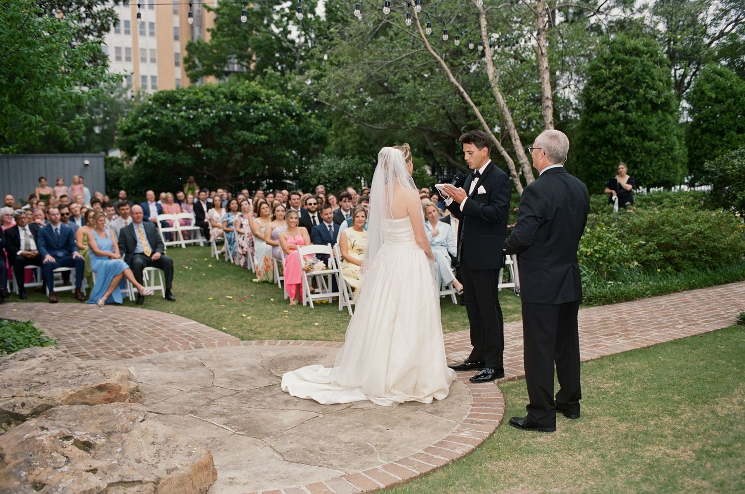 A wedding ceremony takes place outdoors with string lights and trees creating a romantic garden atmosphere.