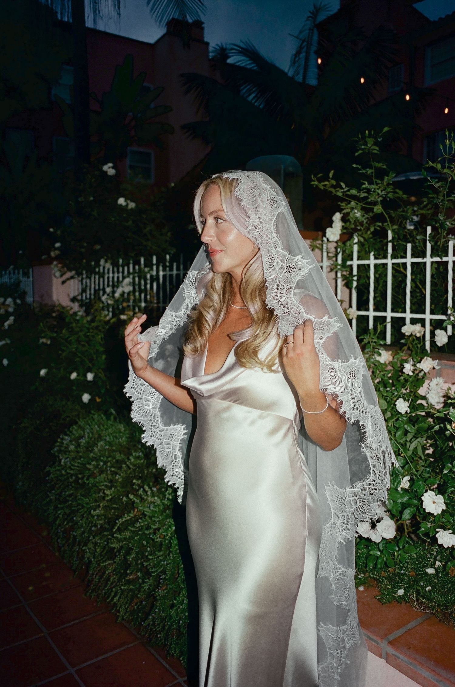 Elegant bride in a silk wedding dress and lace veil poses in a garden at night.