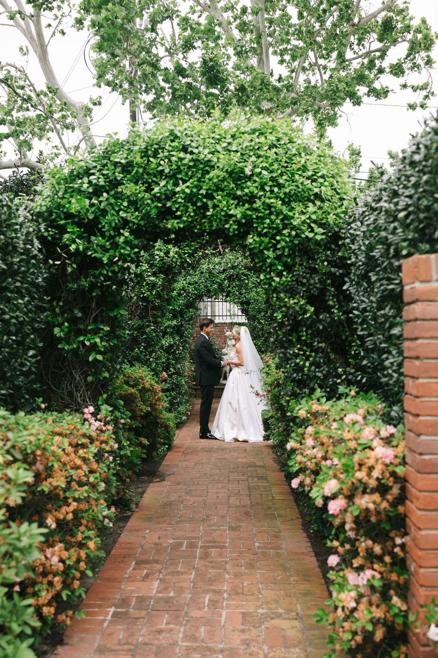 A brick garden pathway lined with green hedges leading to a wedding couple in the distance.