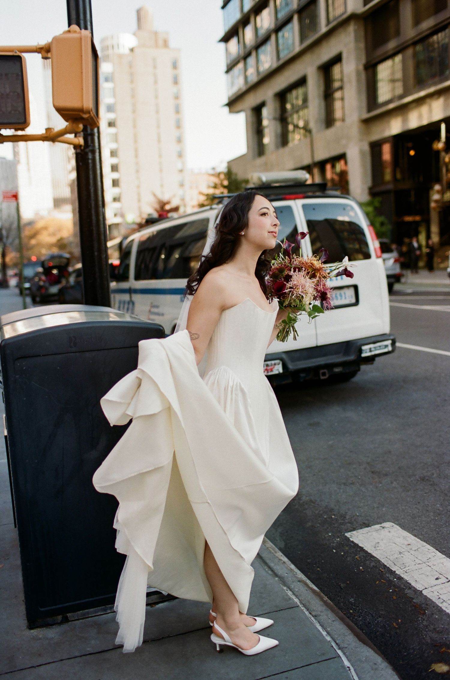 A bride in a flowing white wedding dress holds a bouquet while standing on a city street corner during golden hour.