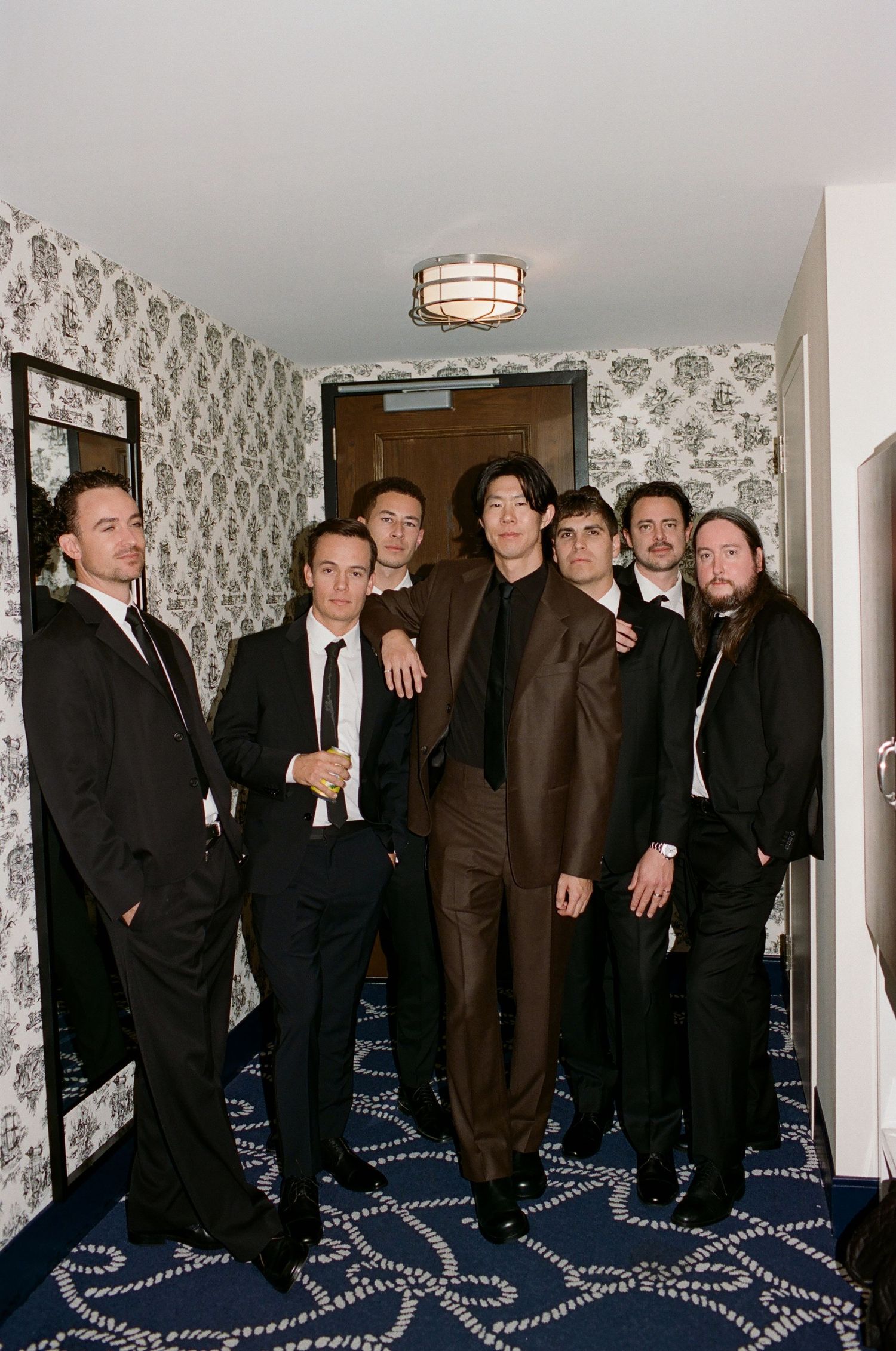 A group of men in formal suits and ties stand together in a hallway with patterned wallpaper and carpeting.