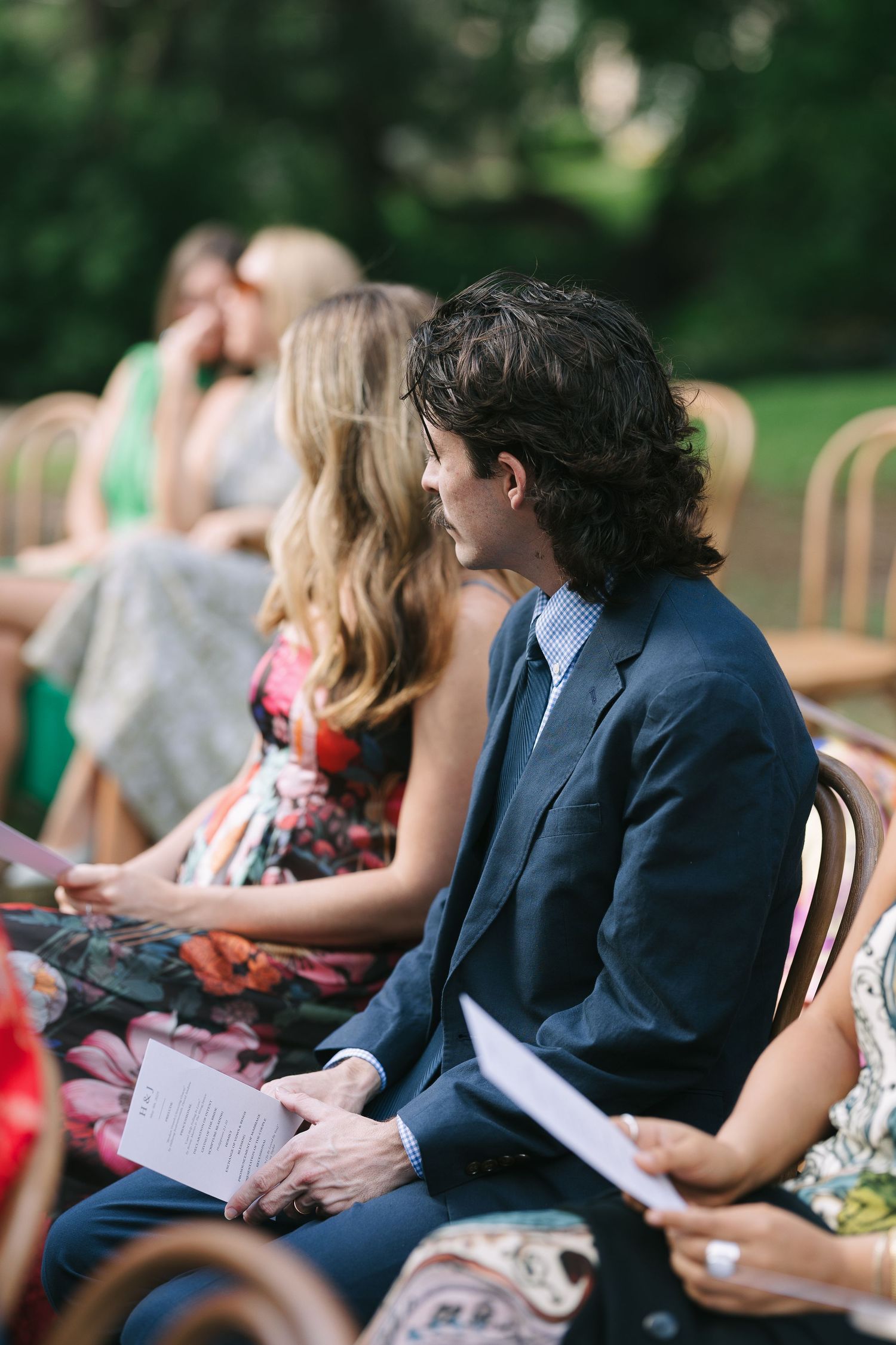 Guests seated in wooden chairs holding ceremony programs at an outdoor event.