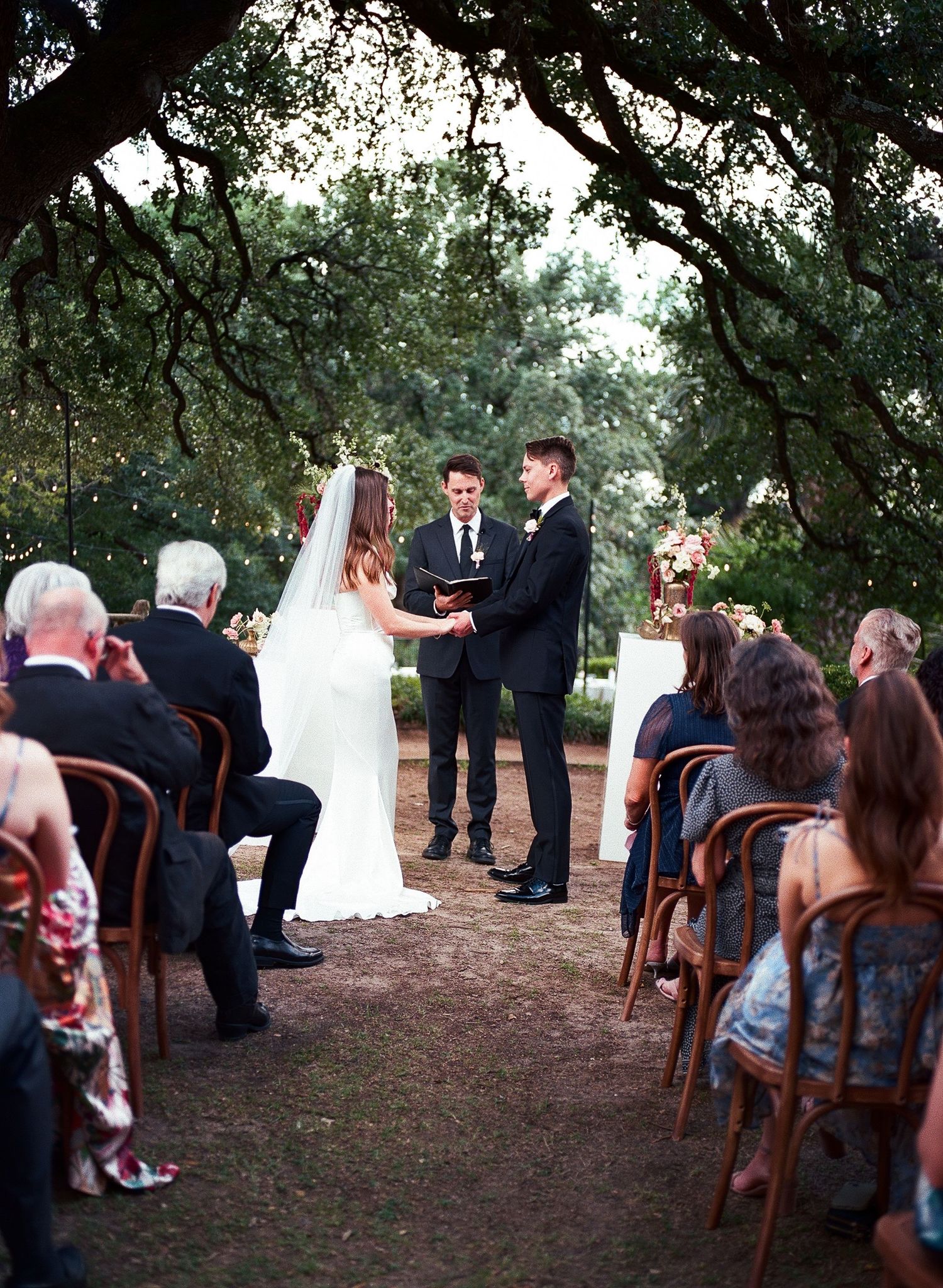 An outdoor wedding ceremony under trees with guests seated on wooden chairs.