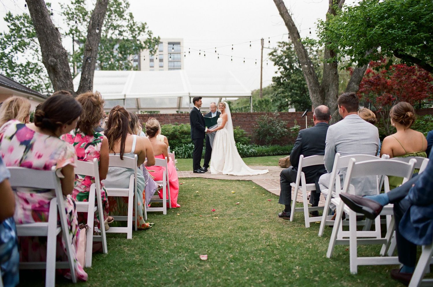 Outdoor wedding ceremony with guests seated on white chairs under string lights.