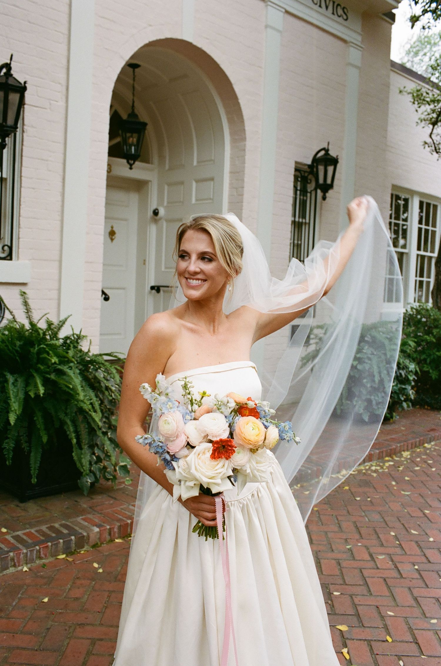 Bride in a strapless white wedding dress holds a peach and white bouquet on a brick patio with white columns.