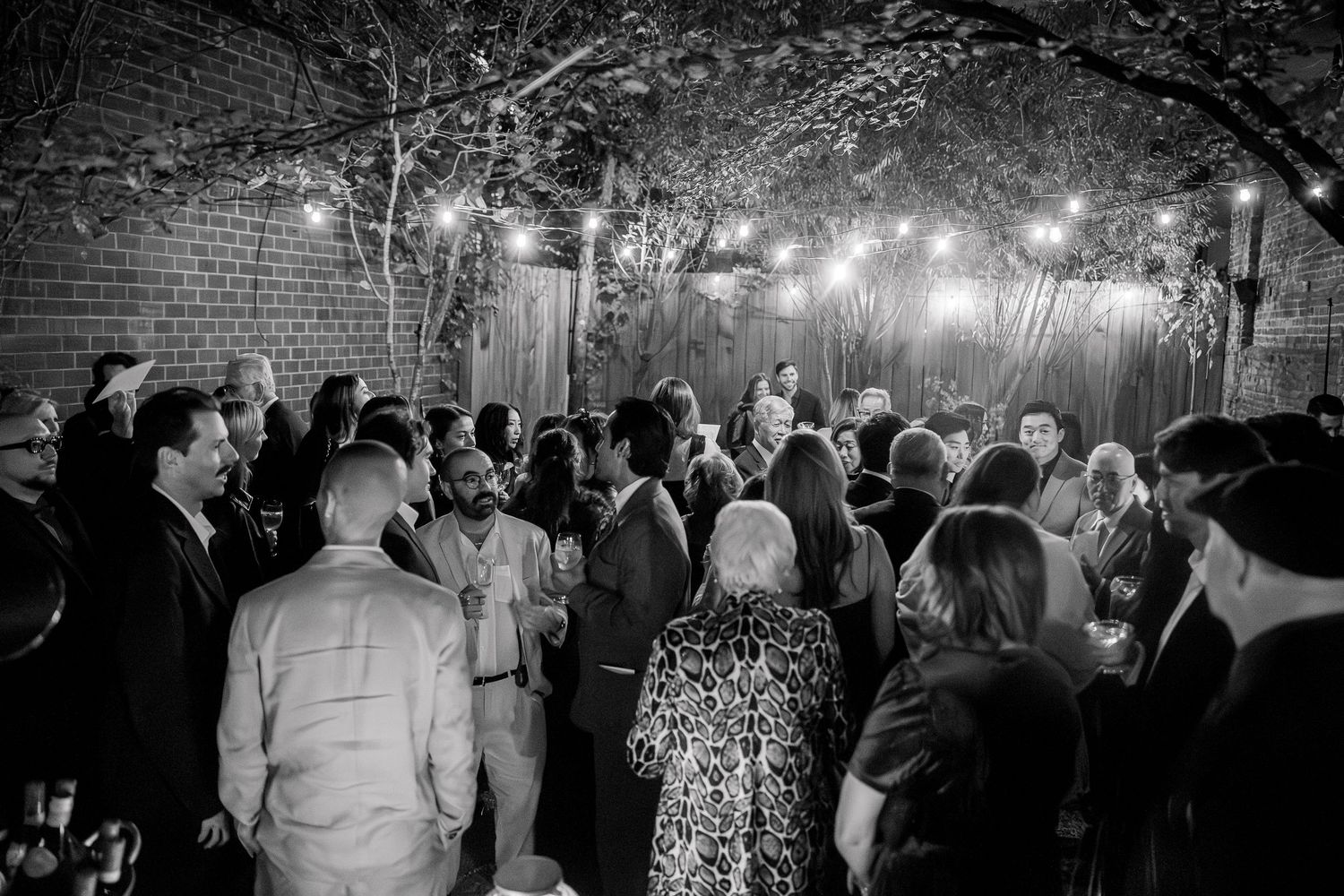 Crowd gathered under string lights and tree branches at an evening outdoor event.