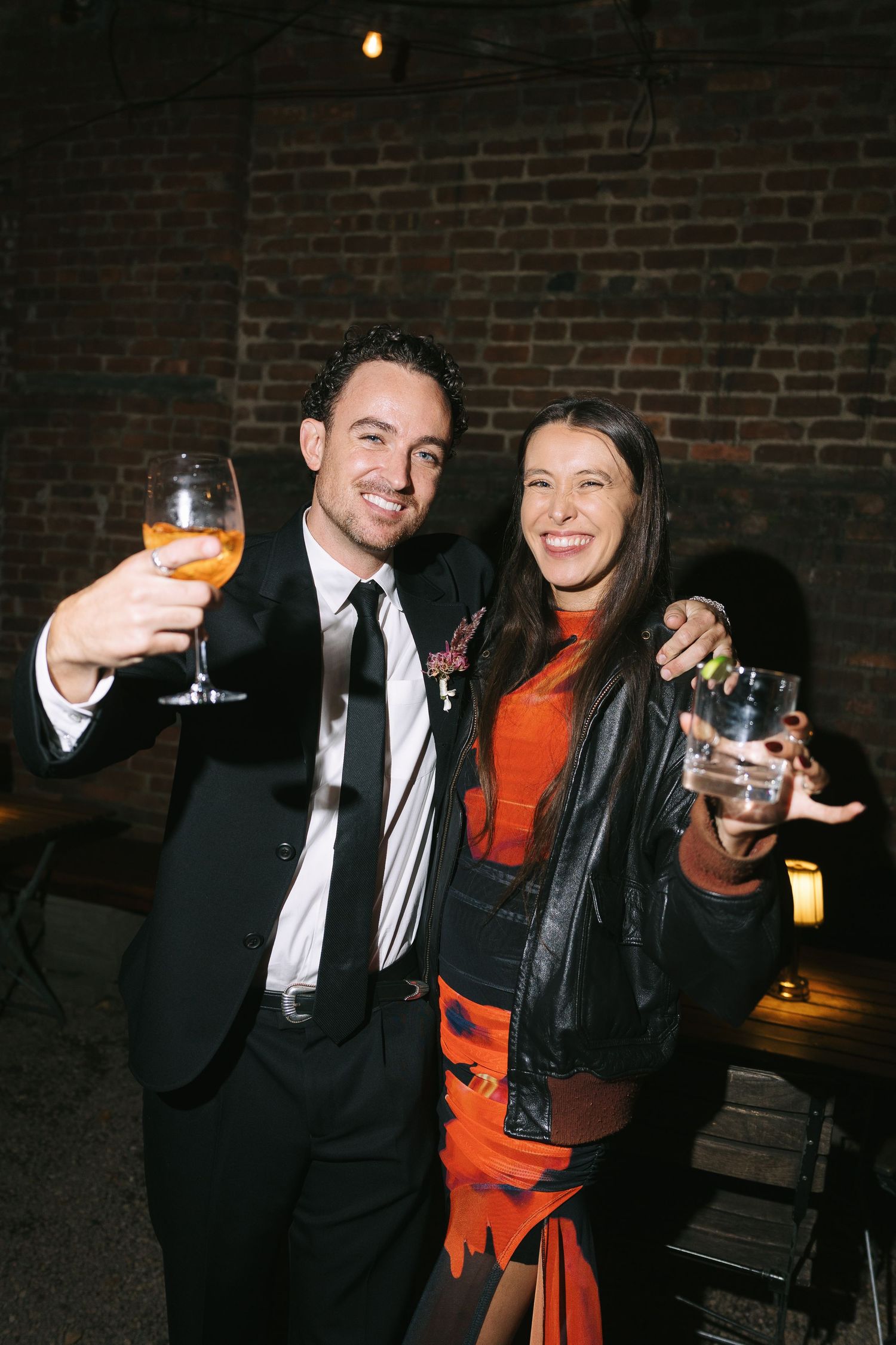 Party guests in formal wear toasting with cocktail glasses against a brick wall backdrop.