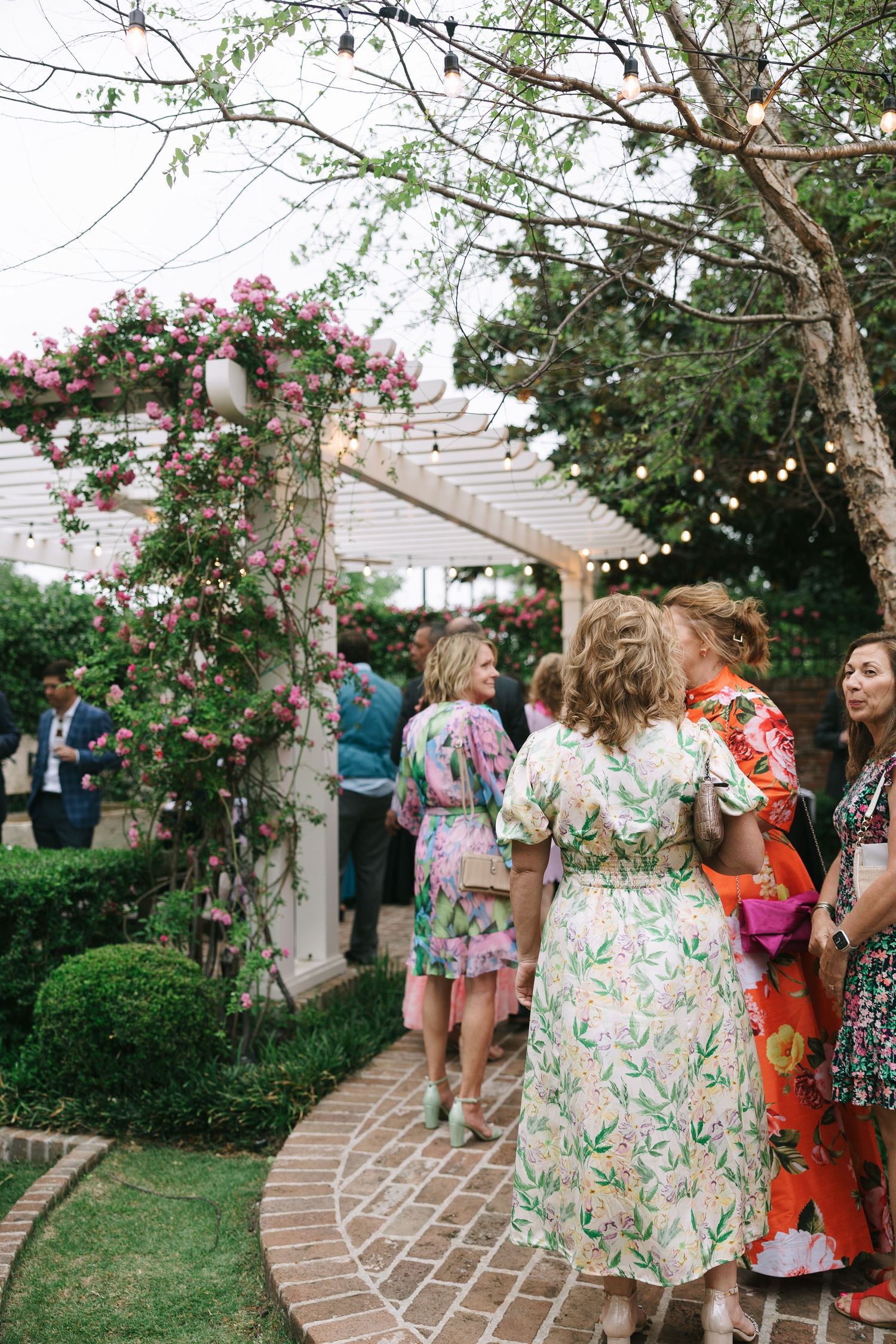 Garden party guests mingling under string lights and flowering trees on a curved brick path.