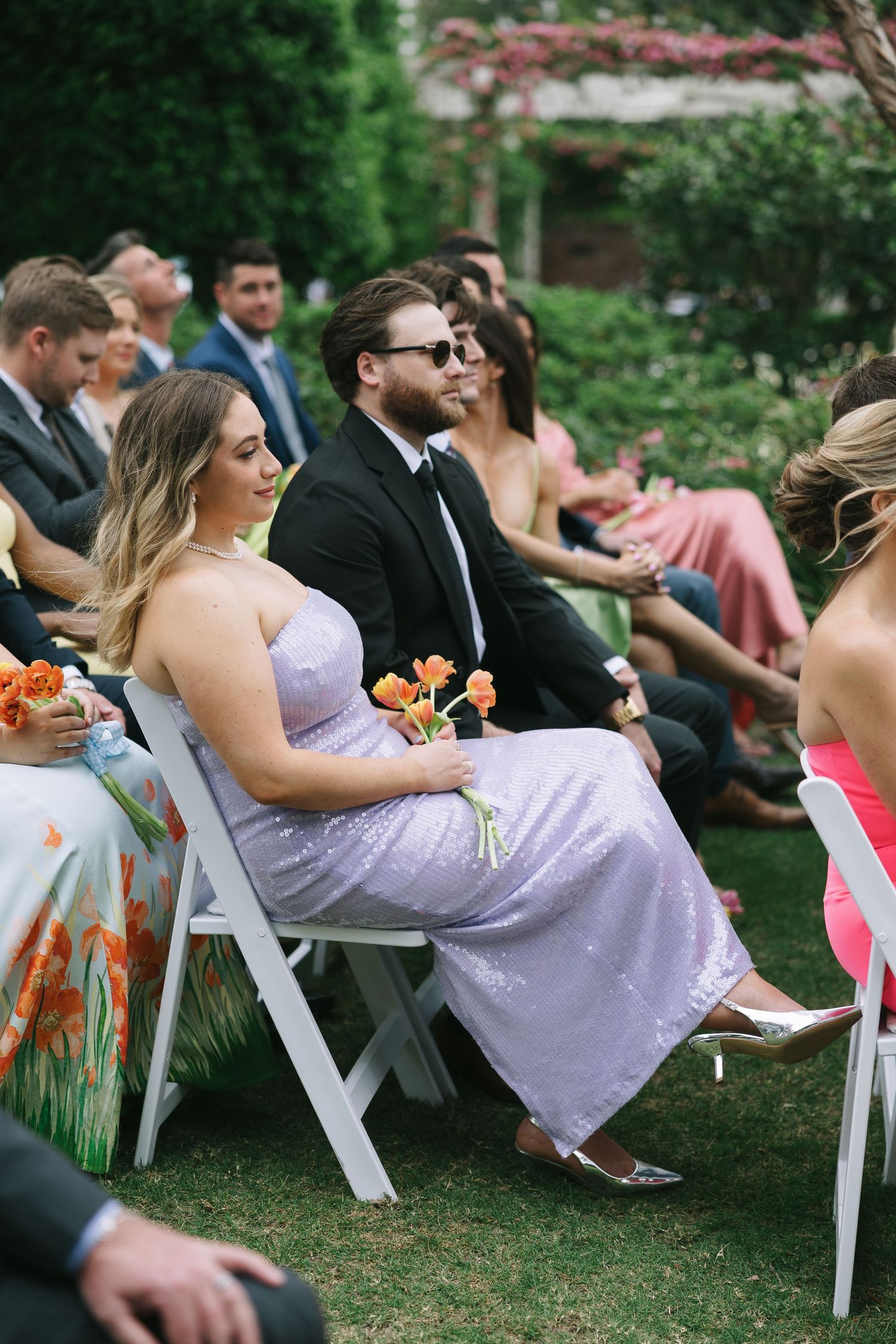 Wedding guests in formal attire and bridesmaids in lavender dresses seated during outdoor ceremony.