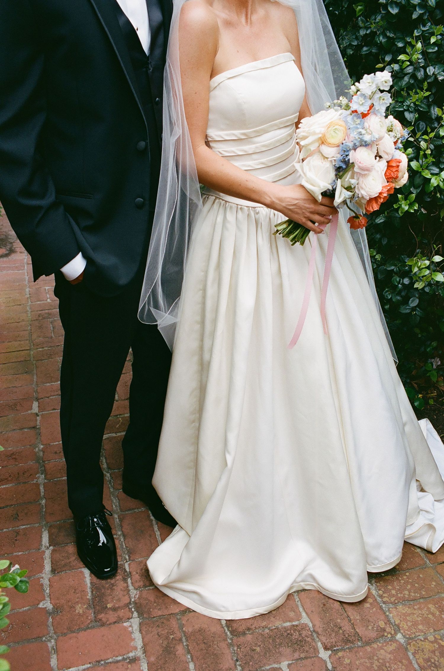 A couple in formal wedding attire poses together on brick pavement.
