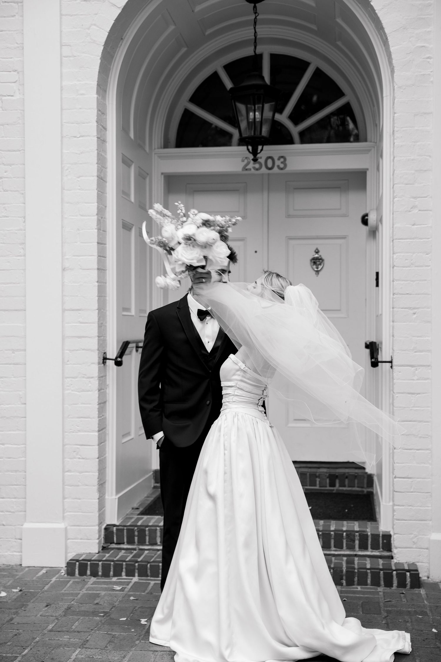 A wedding couple shares a romantic moment in front of a white colonial building with arched doorway.