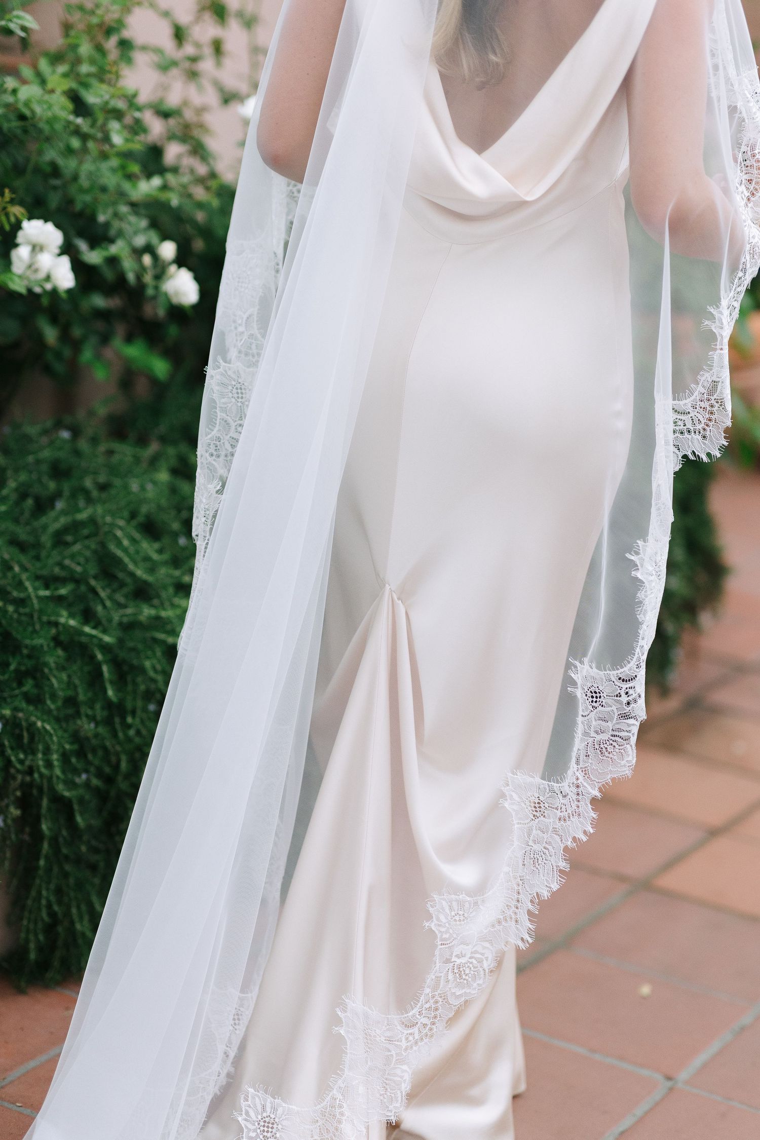 Close-up detail of an elegant white silk wedding dress with delicate lace trim against a garden background.