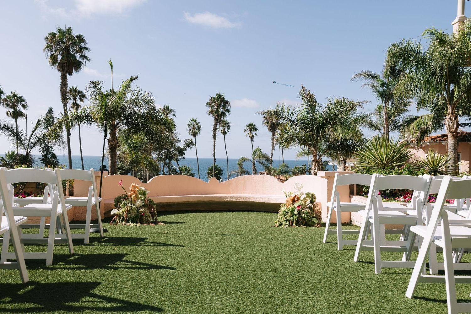 Outdoor wedding venue with white chairs arranged on grass lawn surrounded by palm trees.