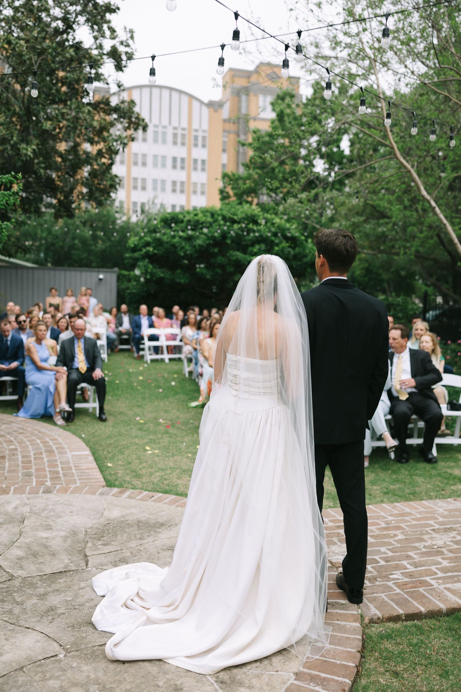 Wedding ceremony on curved brick pathway with string lights and trees overhead.