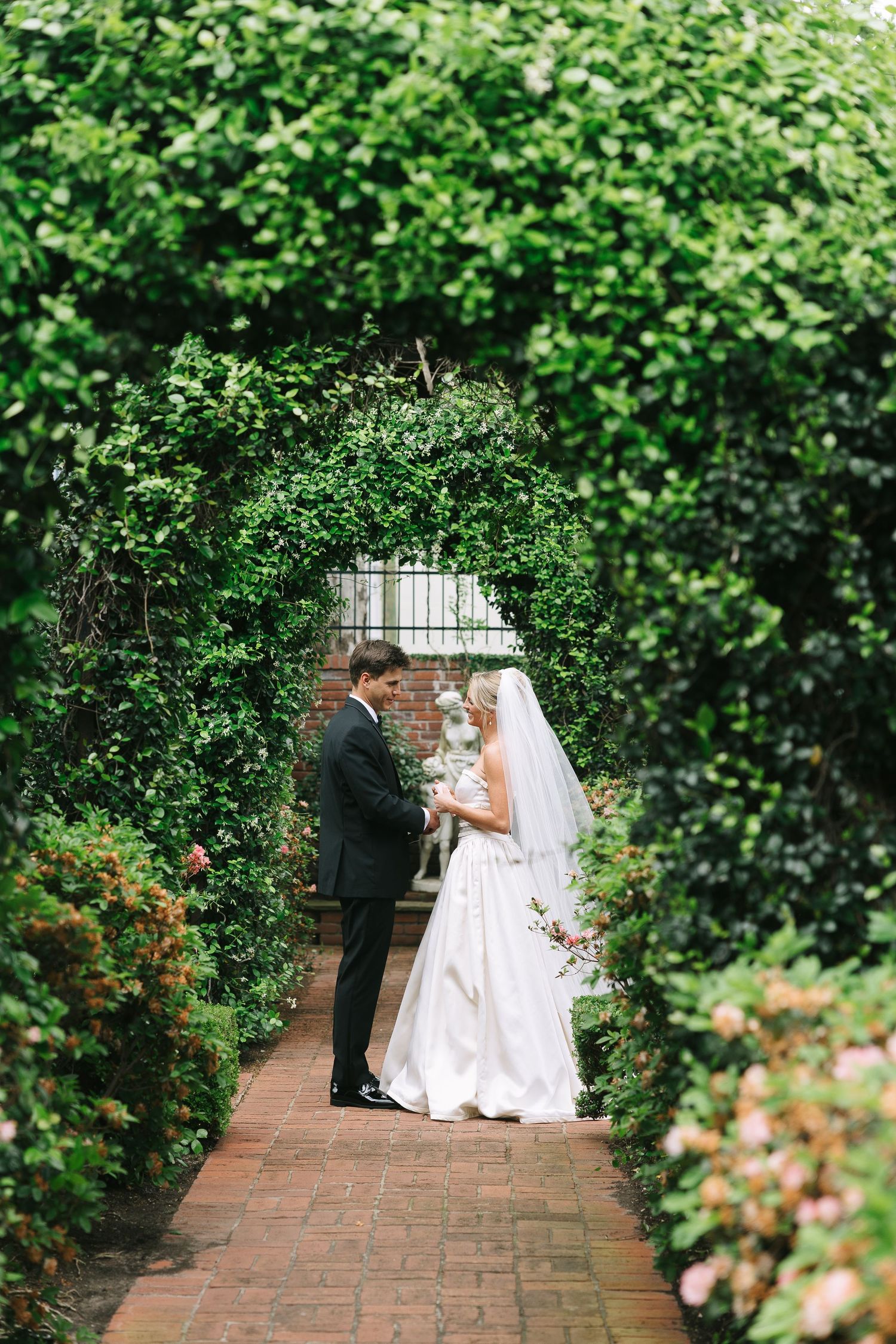 A wedding first look captured in a garden setting with brick pathways and green hedges.
