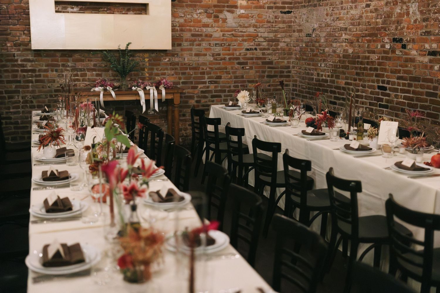 Long dining table setup in a rustic brick-walled venue with floral centerpieces and string lights.