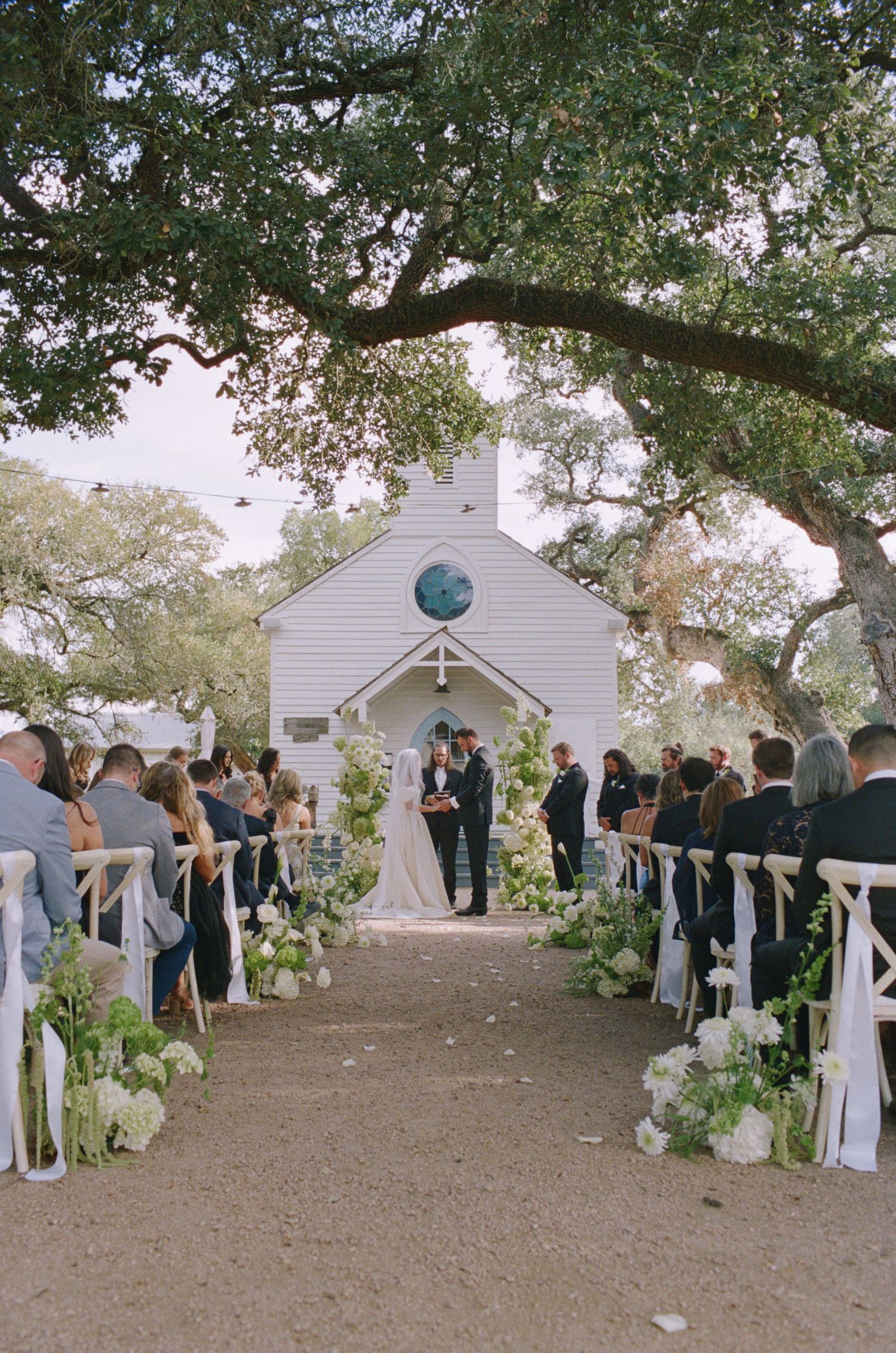 Outdoor wedding ceremony with white chapel, floral-lined aisle, and couple at altar beneath oak trees.