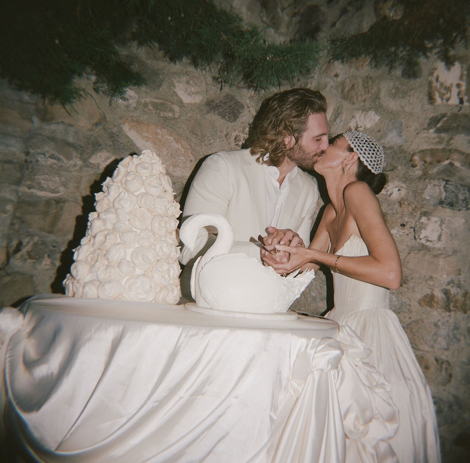 Bride and groom kissing beside elegant white swan wedding cake in rustic stone venue.