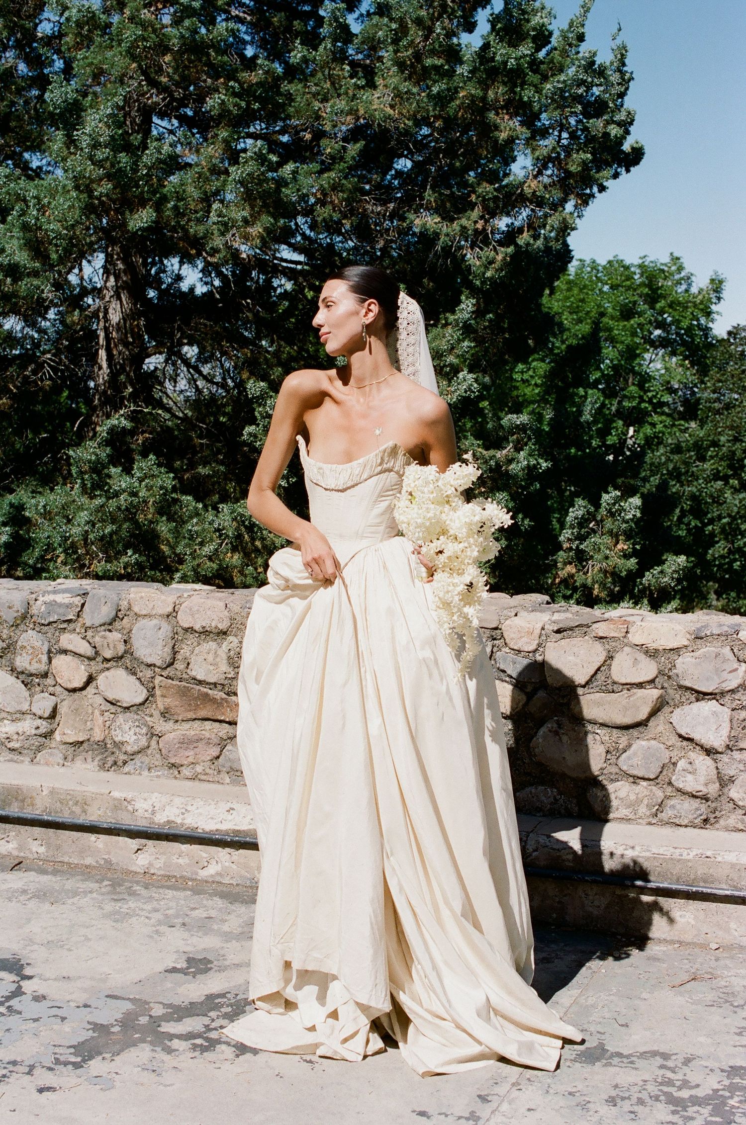 Bride in strapless ivory satin ballgown holding white bouquet, wearing veil, standing by stone wall with trees.