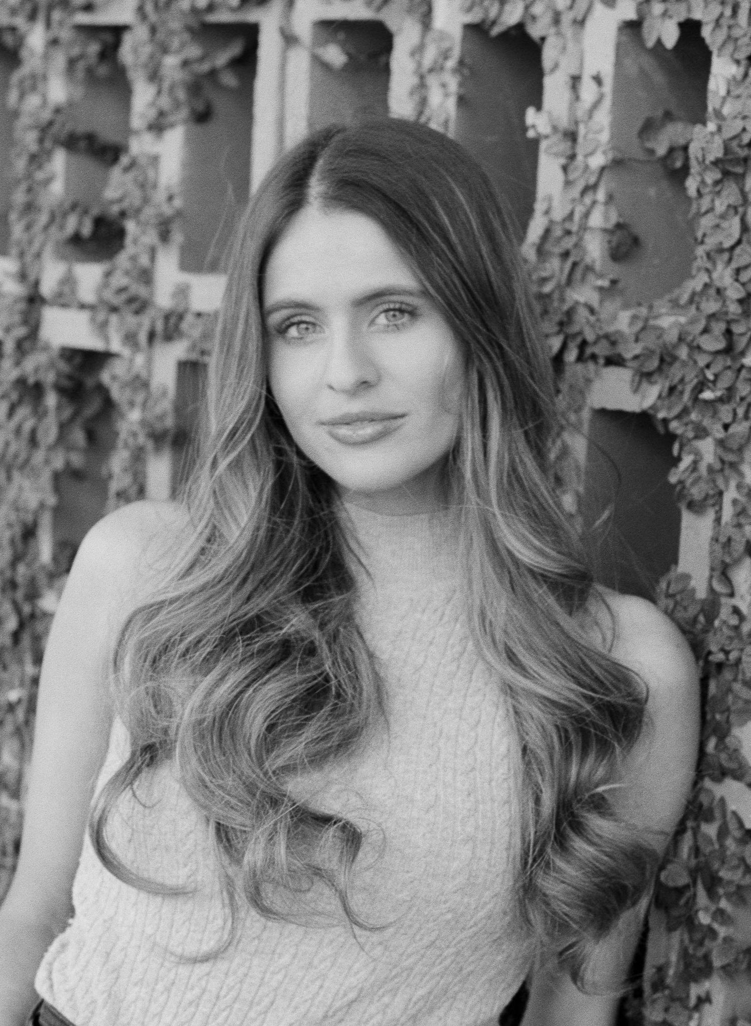 Black and white portrait of a young woman with long wavy hair posing outdoors against a leafy trellis wall.