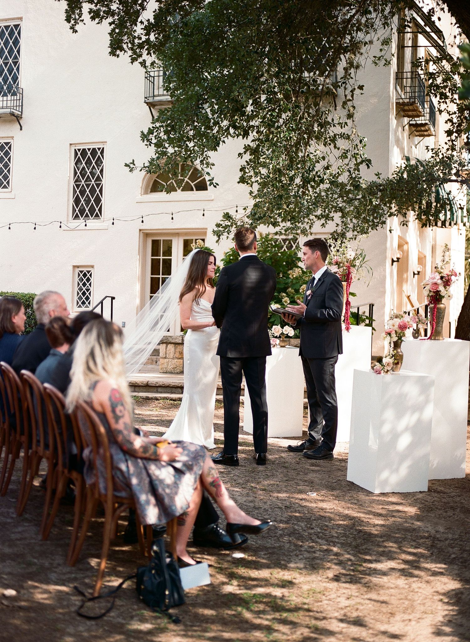 Outdoor wedding ceremony in progress with guests seated on wooden chairs against white building.