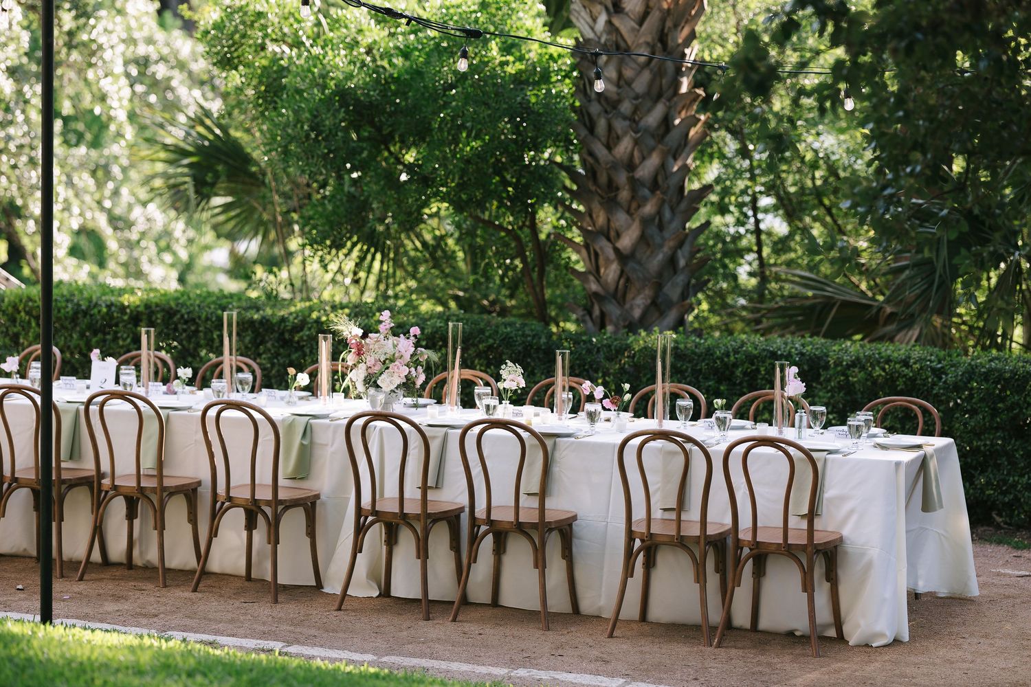 Garden wedding reception setup with white-clothed tables and bentwood chairs under lush trees.
