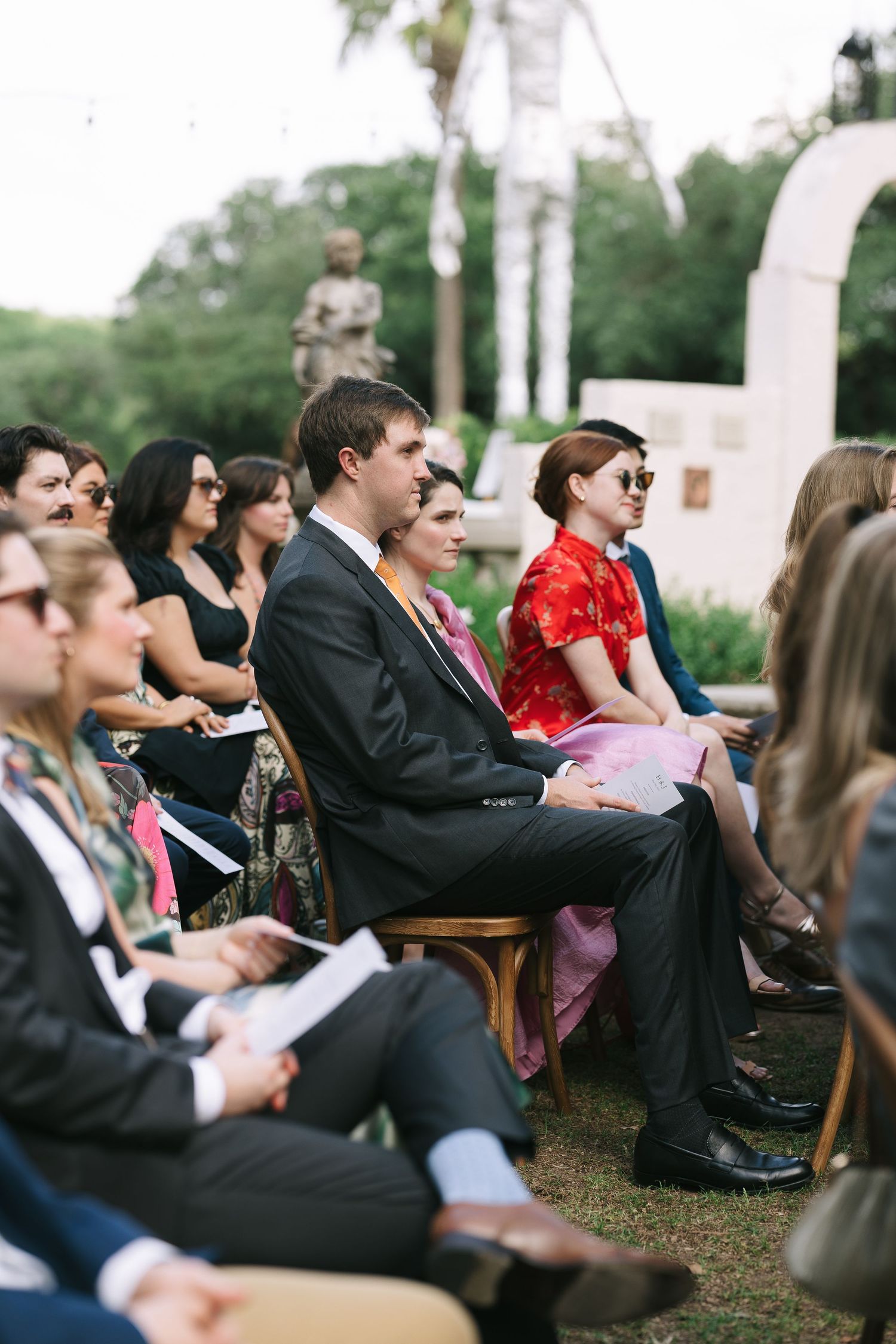 Wedding guests seated in wooden chairs watching an outdoor ceremony under trees.