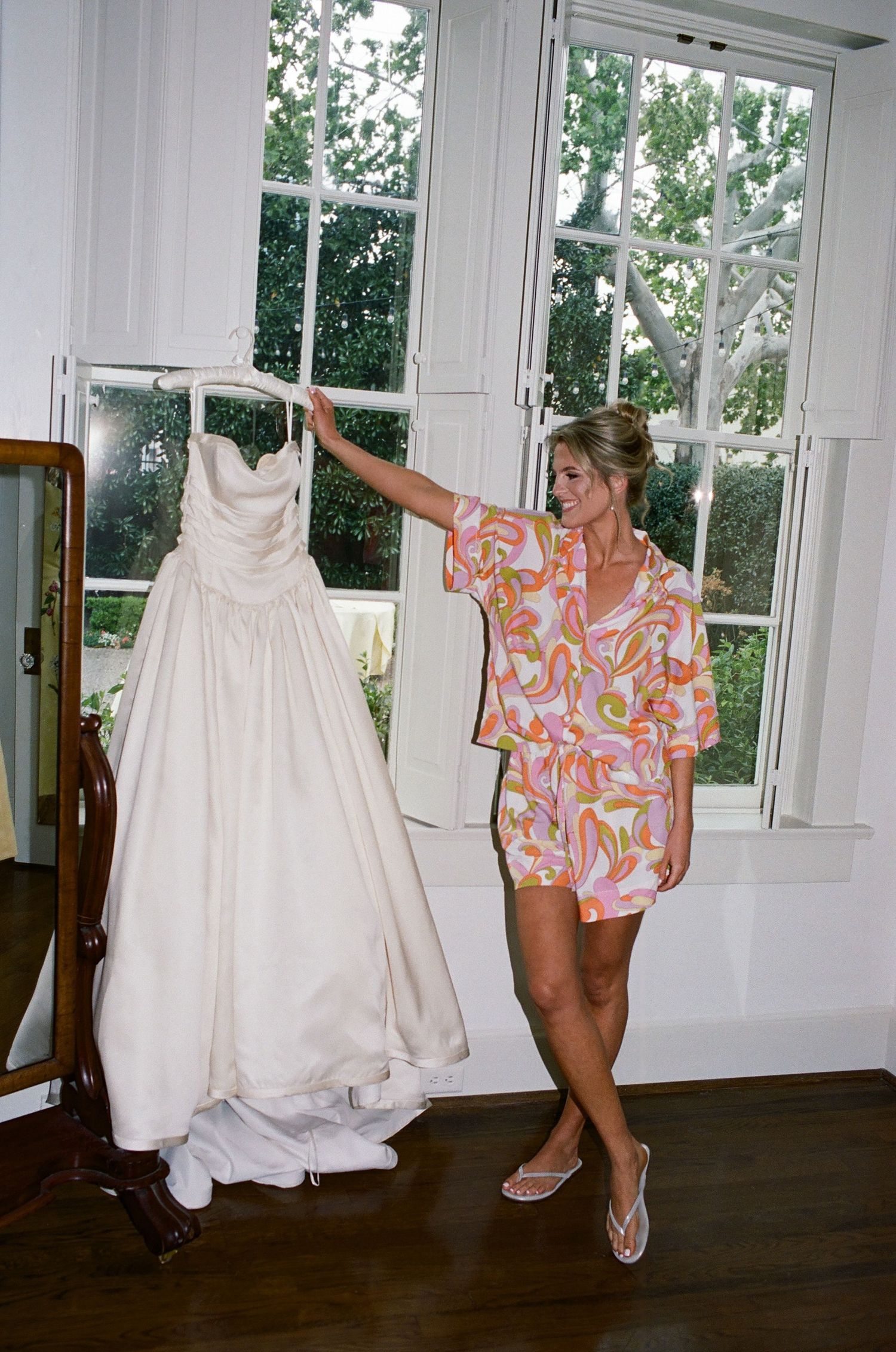 Person in colorful floral dress dancing playfully next to a white wedding gown displayed on a hanger.