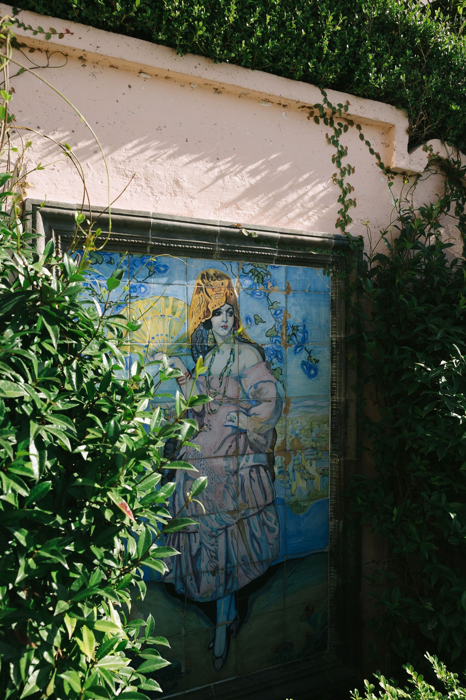 Blue tile mural artwork on white wall surrounded by lush green foliage.
