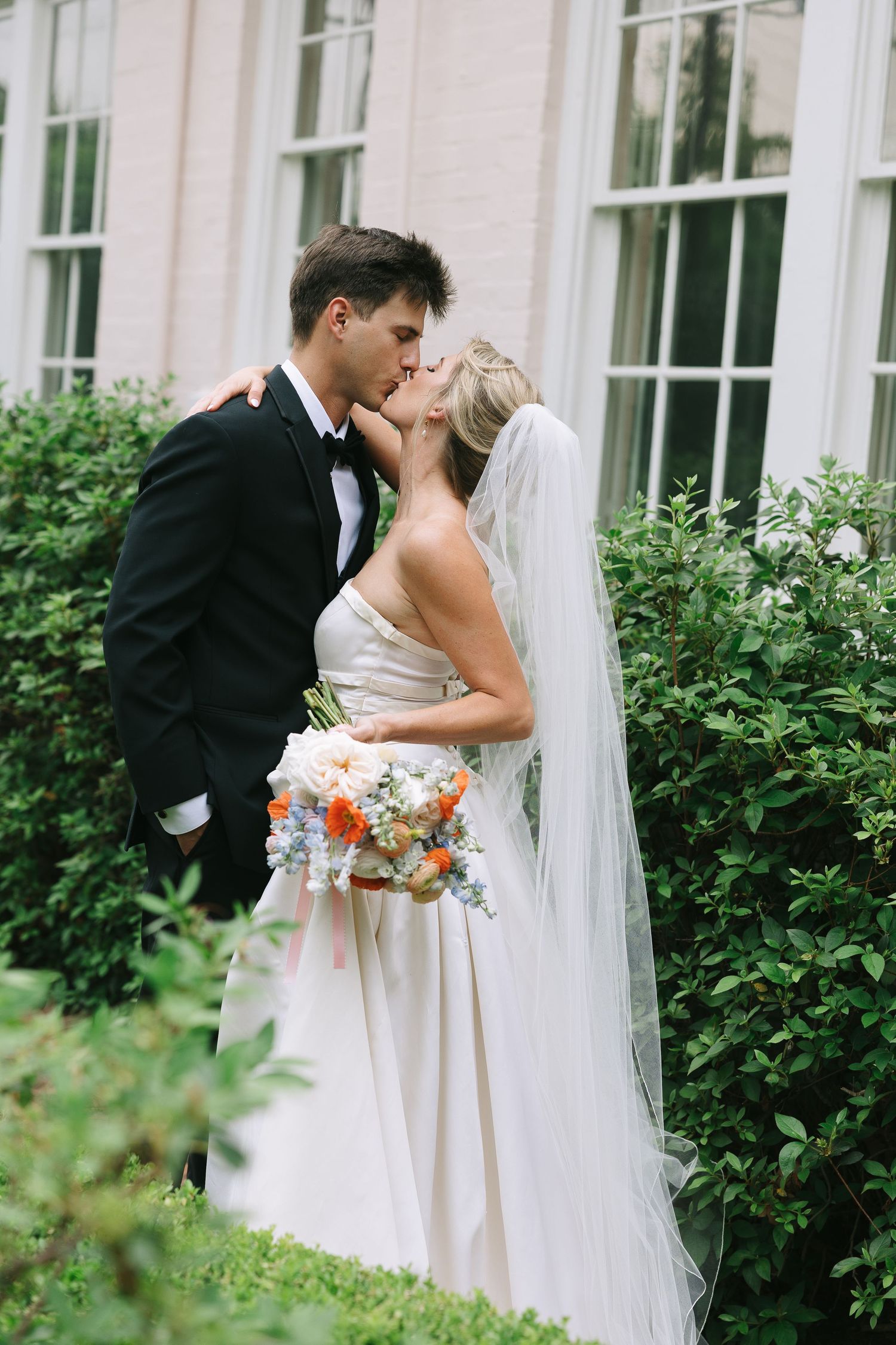 Romantic garden wedding moment captured against white building with windows.