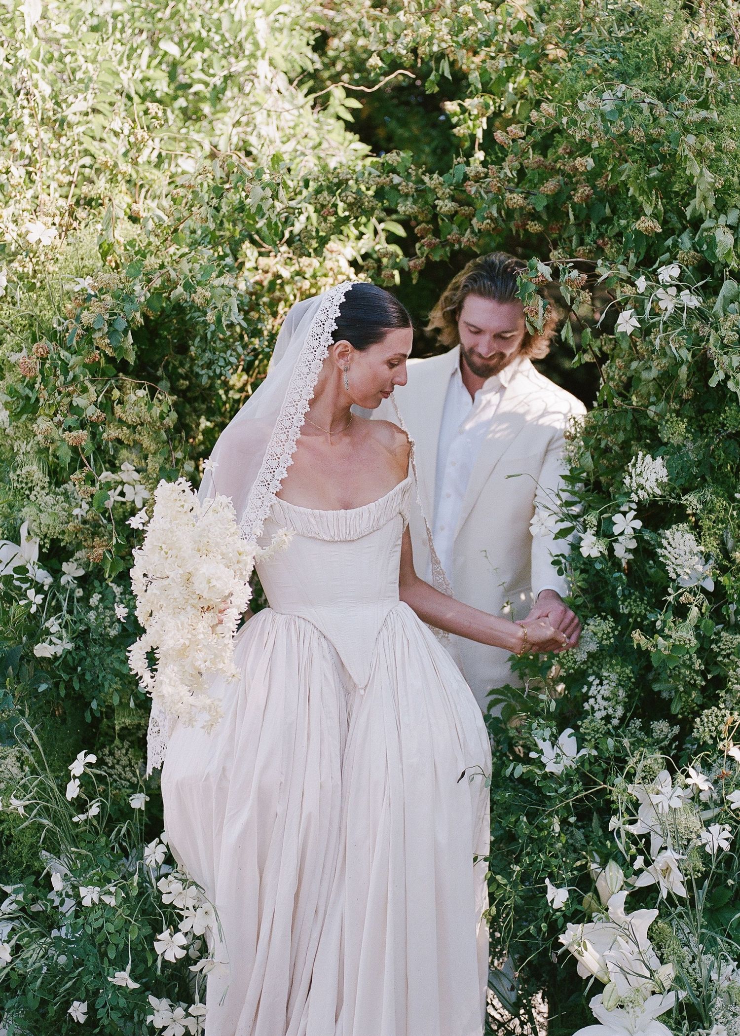 Bride in off-shoulder gown with veil holds white bouquet beside groom in ivory suit among lush green floral arch.