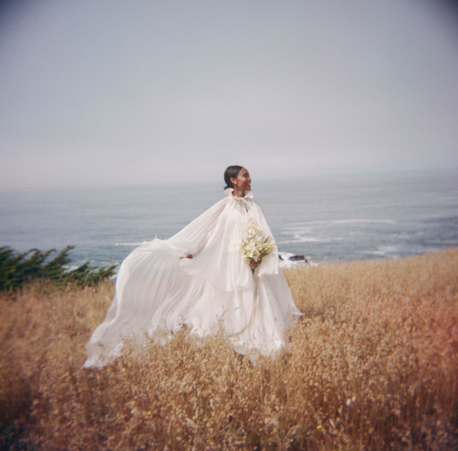 Bride in flowing white cape gown holding bouquet stands in golden field with ocean backdrop on misty day.