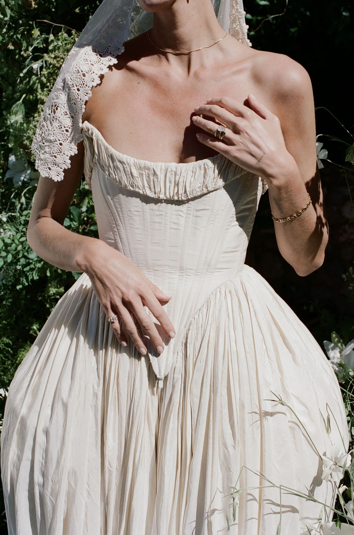 Bride in ivory off-shoulder corset gown with lace detail and veil, posing outdoors among lush greenery.