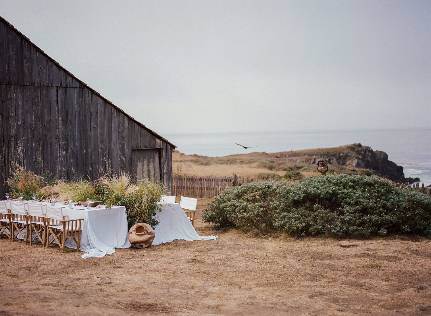Rustic outdoor wedding reception table set near weathered barn with ocean views on California coast.