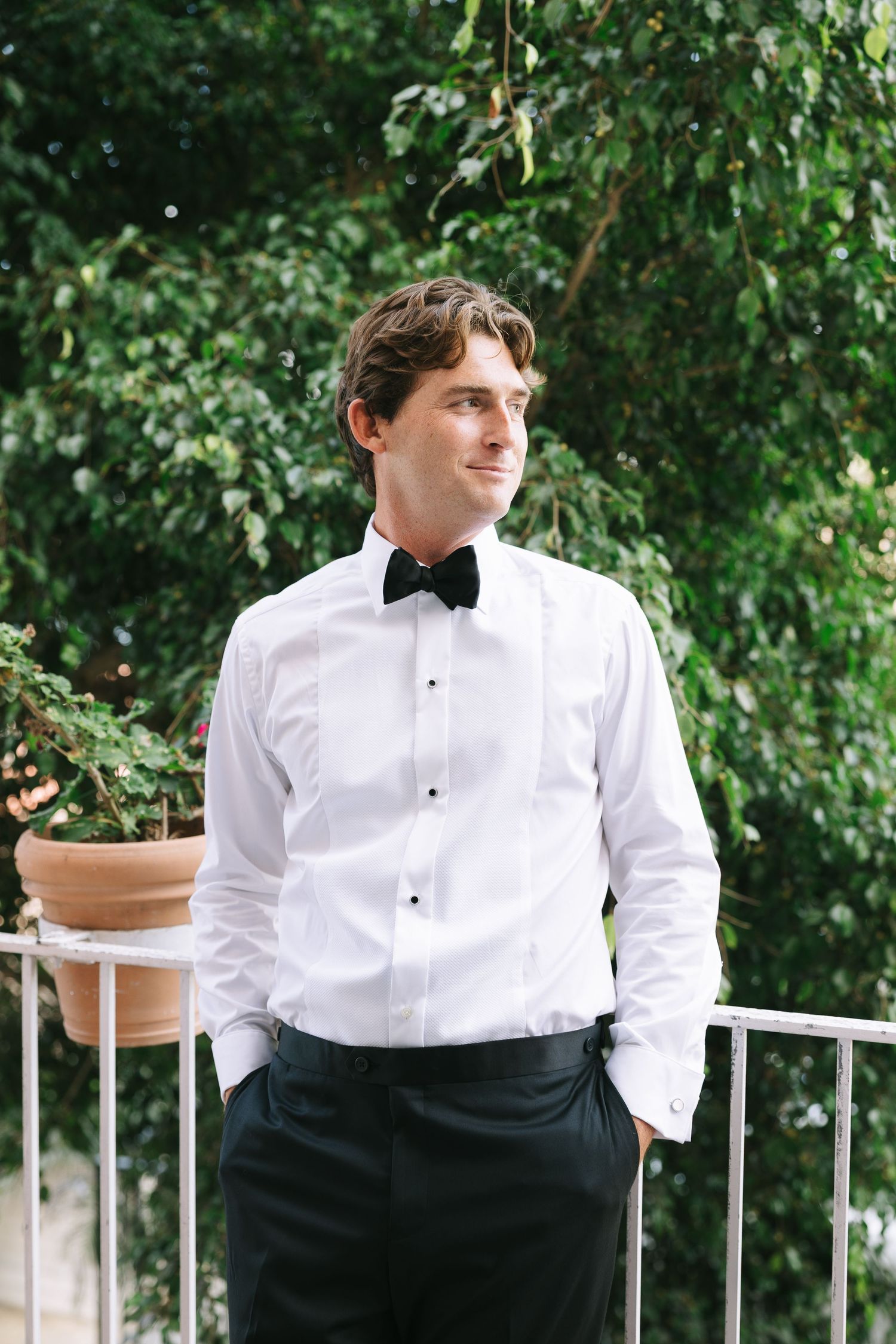 A person in formal attire poses on an outdoor patio with greenery in the background.