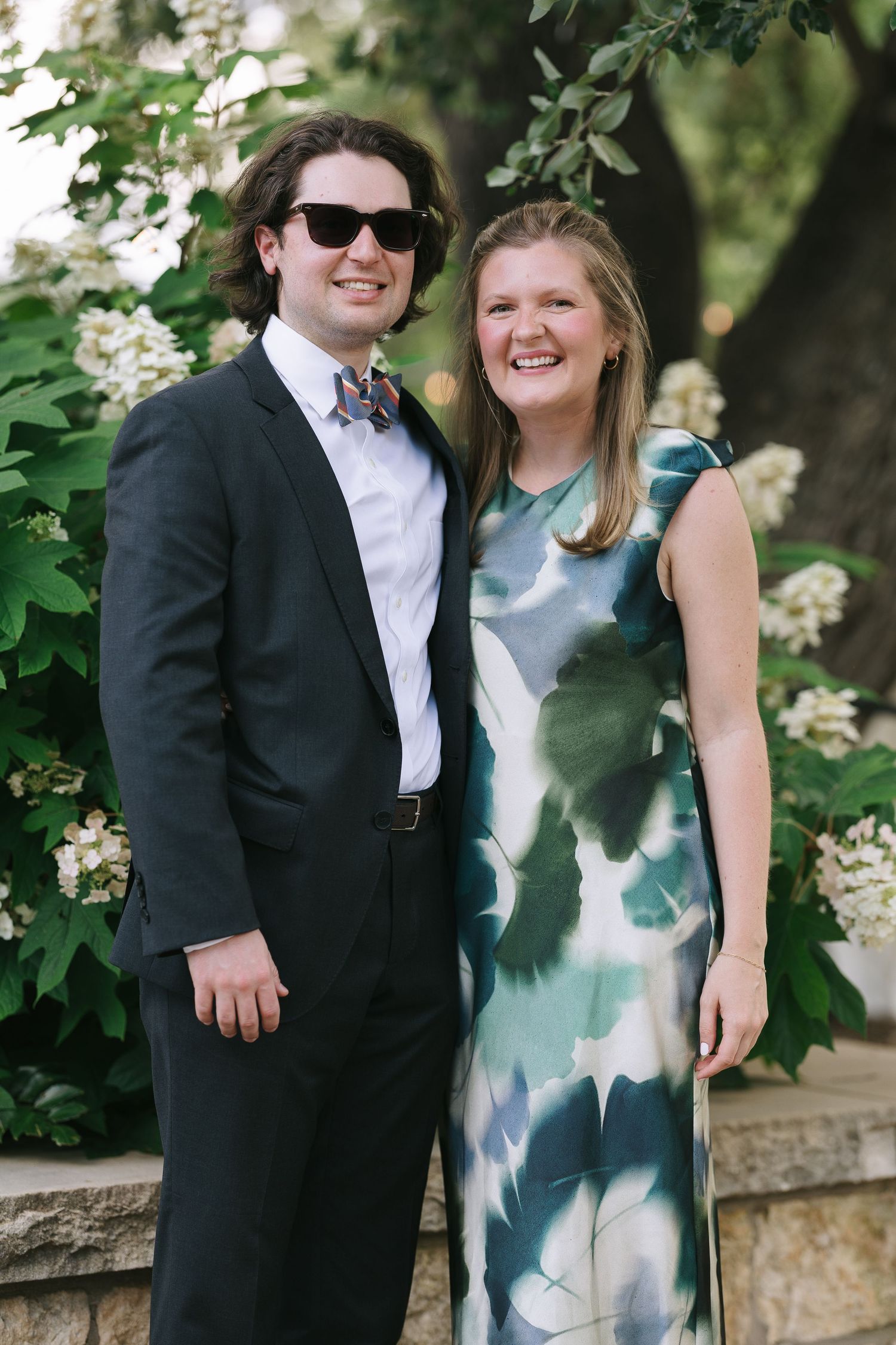 Wedding guests in formal attire pose together on a stone wall under flowering trees.