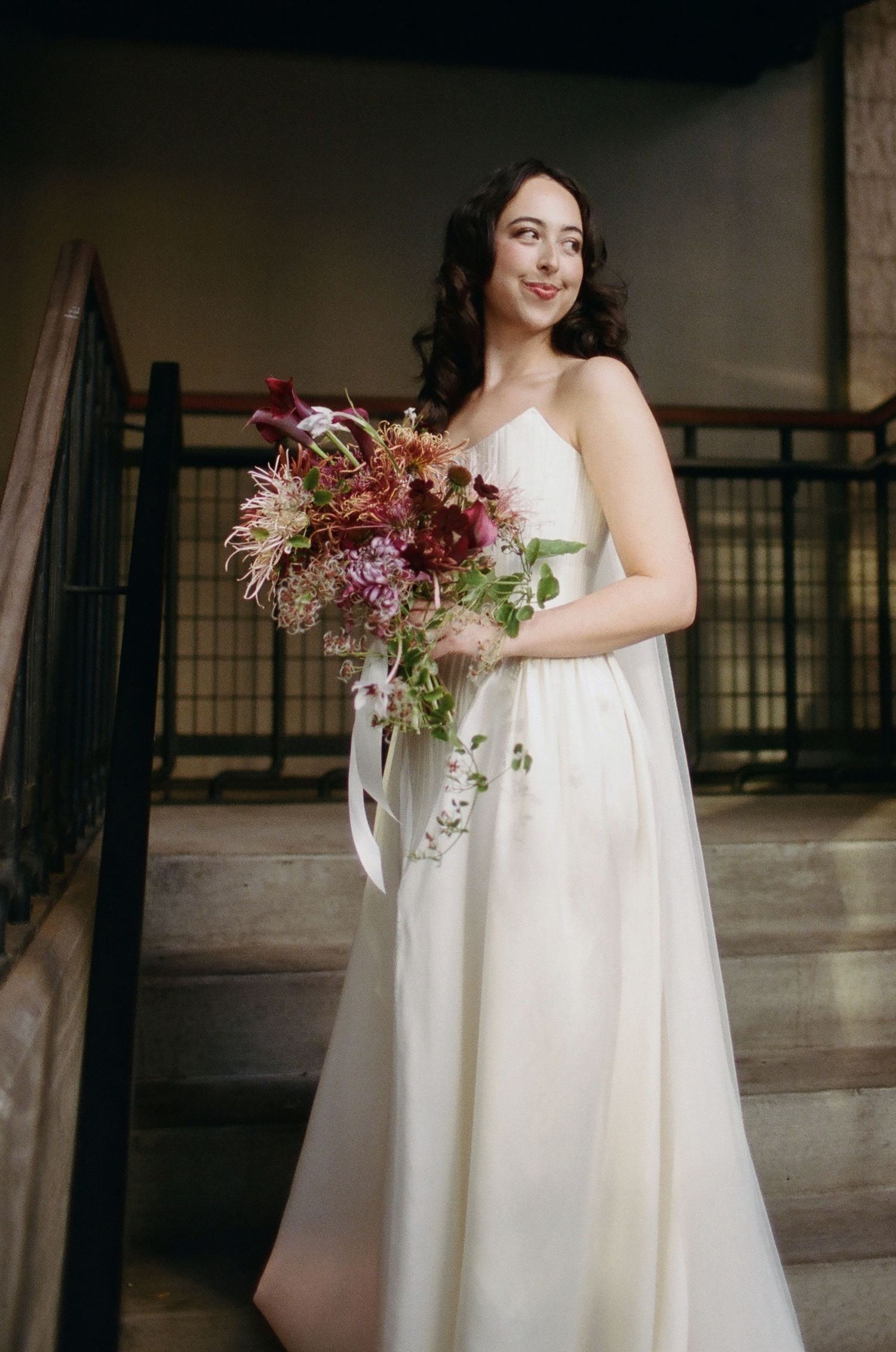 A bride in a flowing white gown holds a cascading burgundy and pink floral bouquet in an industrial venue.