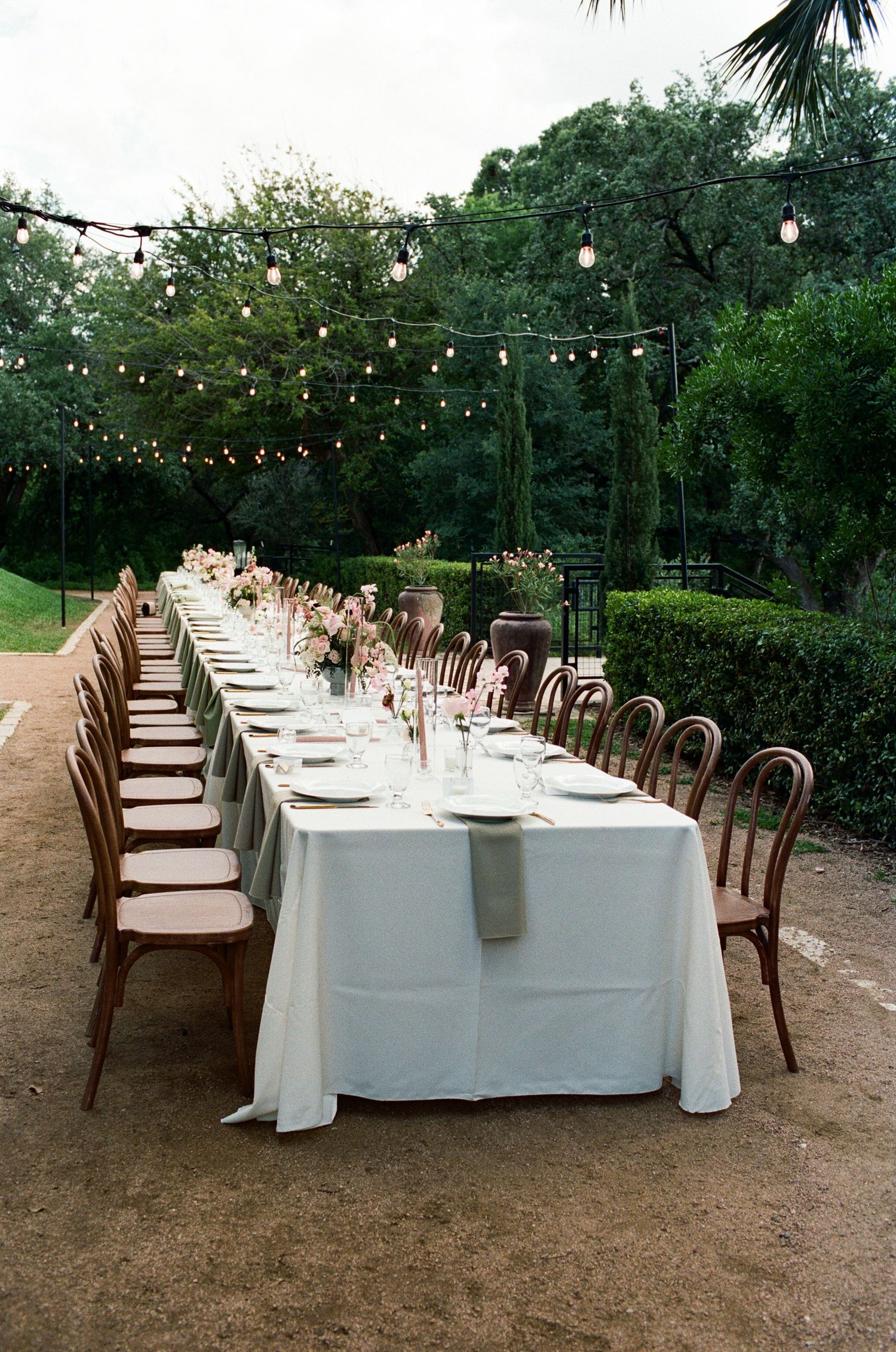 Long dining tables with white linens and wooden chairs set under string lights outdoors.
