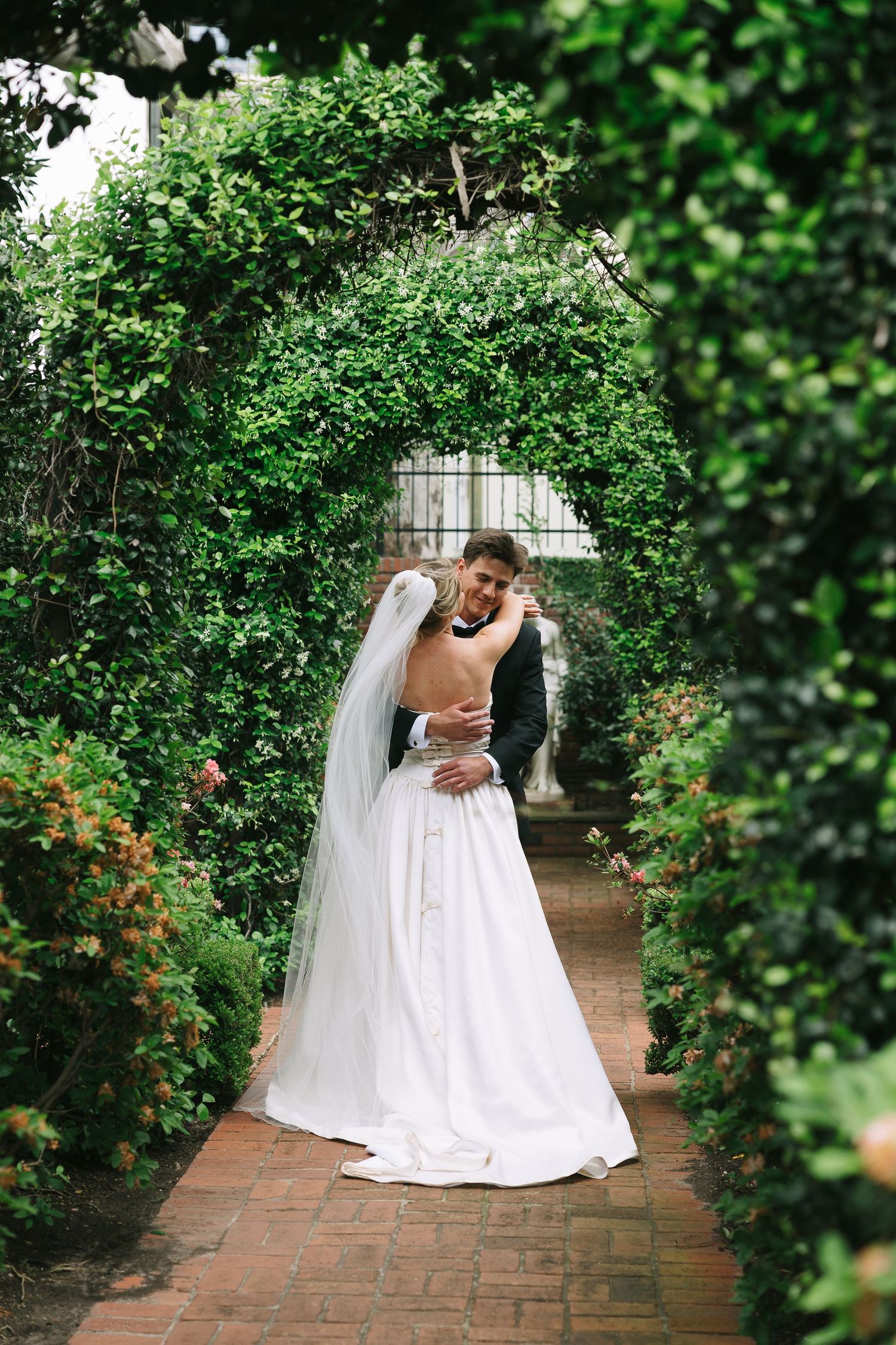 A romantic wedding moment captured on a brick pathway surrounded by blooming garden foliage.