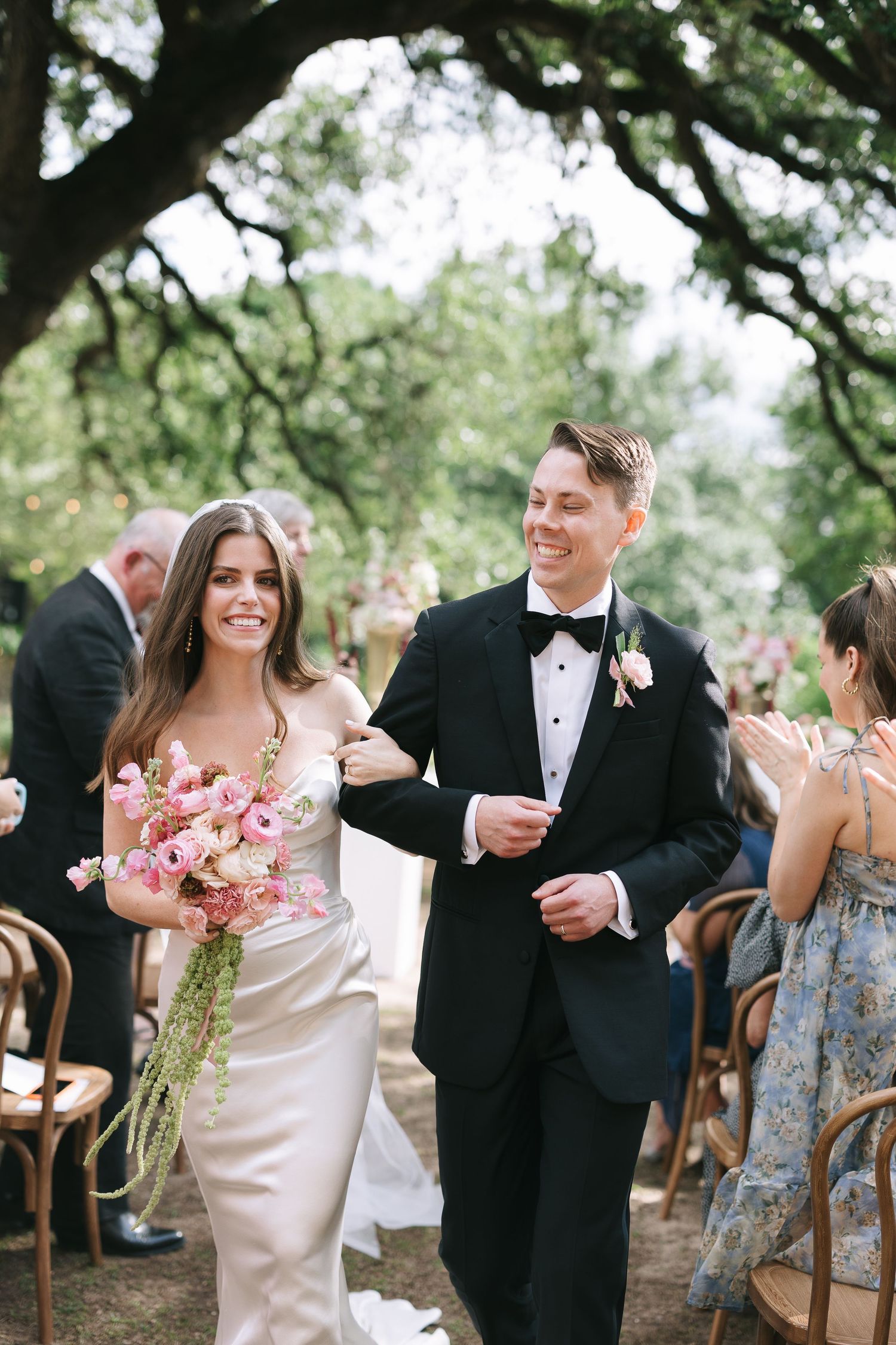 Wedding party members walking down aisle at outdoor ceremony with pink floral bouquets.