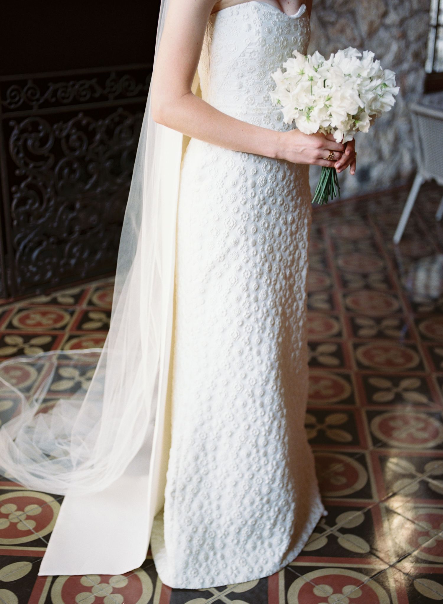 White lace wedding dress detail with floral bouquet against ornate tiled floor.